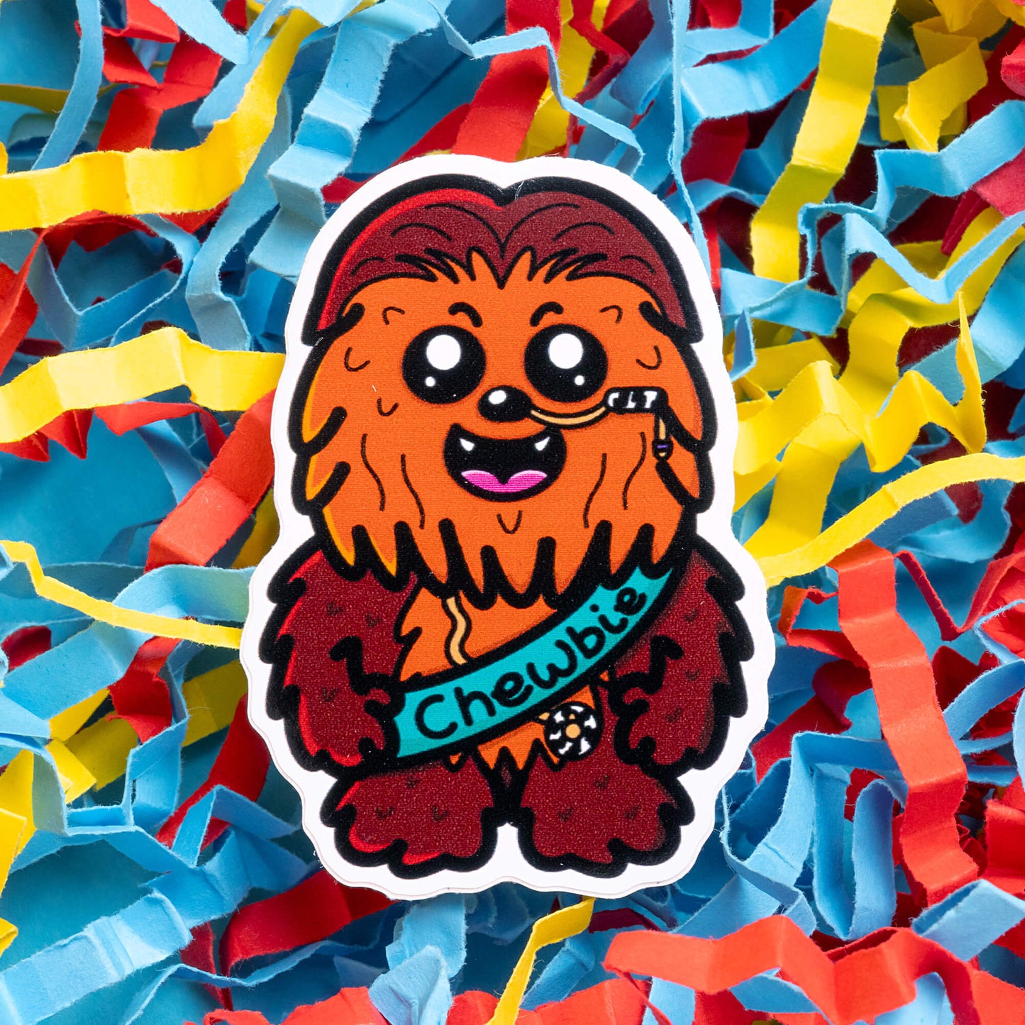 Colourful Innabox sticker of Chewbie, a cartoon character resembling Chewbacca, wearing a feeding tube. Set against a backdrop of red, blue, and yellow shredded paper, this sticker promotes awareness for hidden disabilities and is part of Innabox’s collection of uplifting products for people with chronic illnesses.