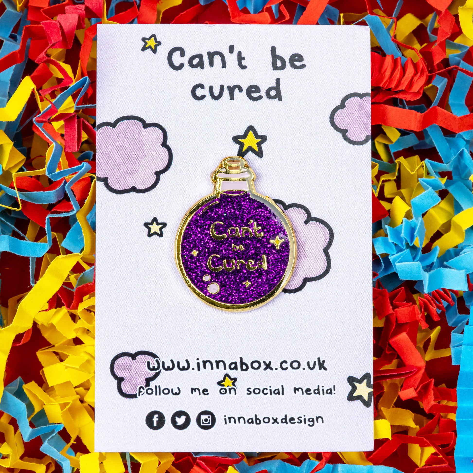 The Can't Be Cured Potion Bottle Enamel Pin on a lilac backing card with purple clouds and yellow stars, with can't be cured written at the top and innabox social media handles written at the bottom both in black, on a card confetti background. The pin is a gold outlined circular potion bottle with a glittery purple middle with two pastel pink bubbles, gold sparkles and gold text that reads 'can't be cured'. The design was inspired by chronic illnesses.