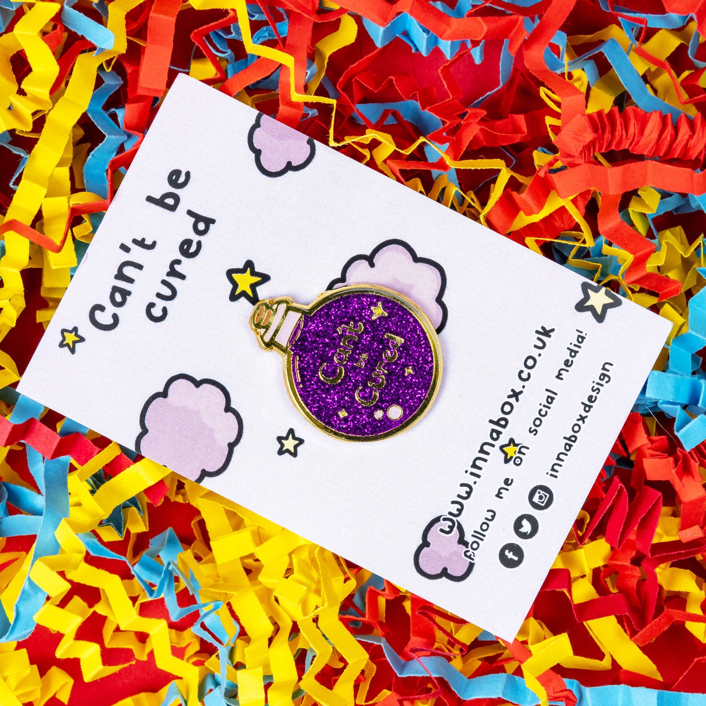 The Can't Be Cured Potion Bottle Enamel Pin on a lilac backing card with purple clouds and yellow stars, with can't be cured written at the top and innabox social media handles written at the bottom both in black, on a card confetti background. The pin is a gold outlined circular potion bottle with a glittery purple middle with two pastel pink bubbles, gold sparkles and gold text that reads 'can't be cured'. The design was inspired by chronic illnesses.