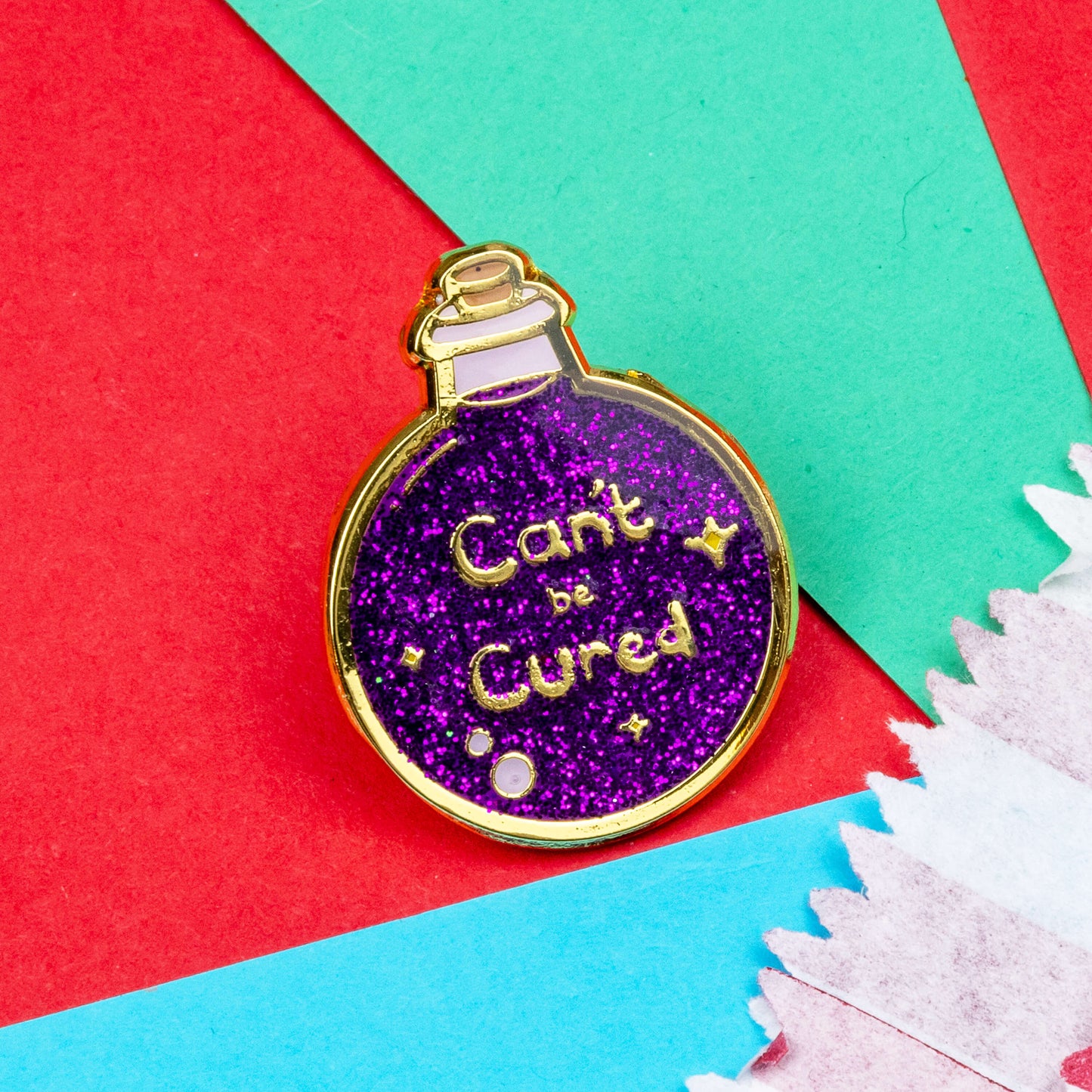 The Can't Be Cured Potion Bottle Enamel Pin on a blue, red and teal background. A gold outlined circular potion bottle with a glittery purple middle with two pastel pink bubbles, gold sparkles and gold text that reads 'can't be cured'. The design was inspired by chronic illnesses.