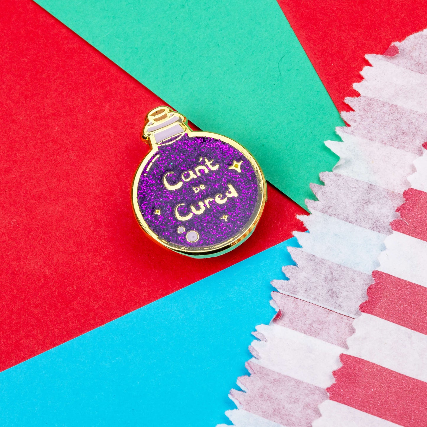 The Can't Be Cured Potion Bottle Enamel Pin on a blue, red and teal background. A gold outlined circular potion bottle with a glittery purple middle with two pastel pink bubbles, gold sparkles and gold text that reads 'can't be cured'. The design was inspired by chronic illnesses.