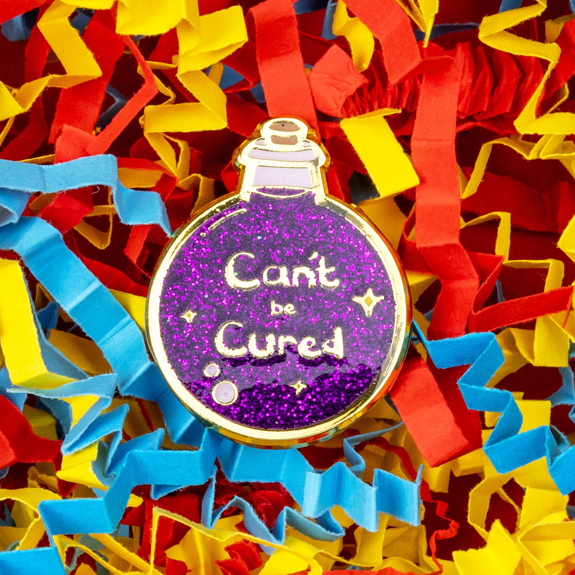 The Can't Be Cured Potion Bottle Enamel Pin on a blue, red and yellow card confetti background. A gold outlined circular potion bottle with a glittery purple middle with two pastel pink bubbles, gold sparkles and gold text that reads 'can't be cured'. The design was inspired by chronic illnesses.