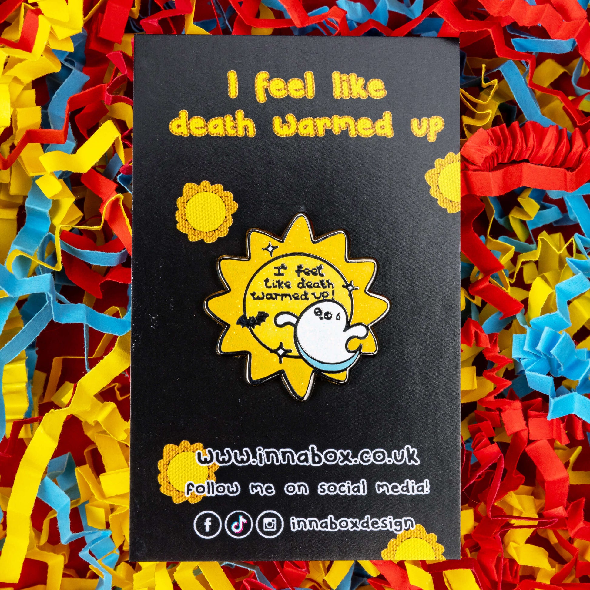 Death Warmed Up Halloween Pin on black backing card with yellow suns on on a red, blue and yellow coloured card confetti background. The enamel pin is a glittery yellow sun pin with 'I feel like death warmed up!' in the middle with white sparkles. There is a little black bat and a cute white ghost with a smile and a sweat droplet on its forehead. Hand drawn to raise awareness for chronic illness
