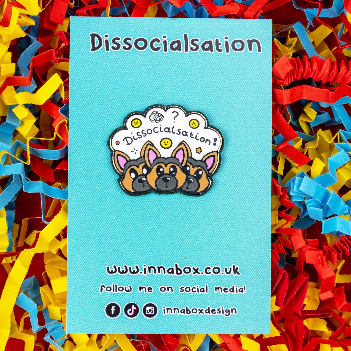The Dissocialsation Enamel Pin - Dissociation on blue backing card on a red, blue and yellow card confetti background. Three confused brown and black Alsatian dog heads with their ears perked up, above them is a white cloud with sad and happy yellow faces, sparkles, question marks and black text reading 'dissocialsation!'. The design is raising awareness for dissociation.