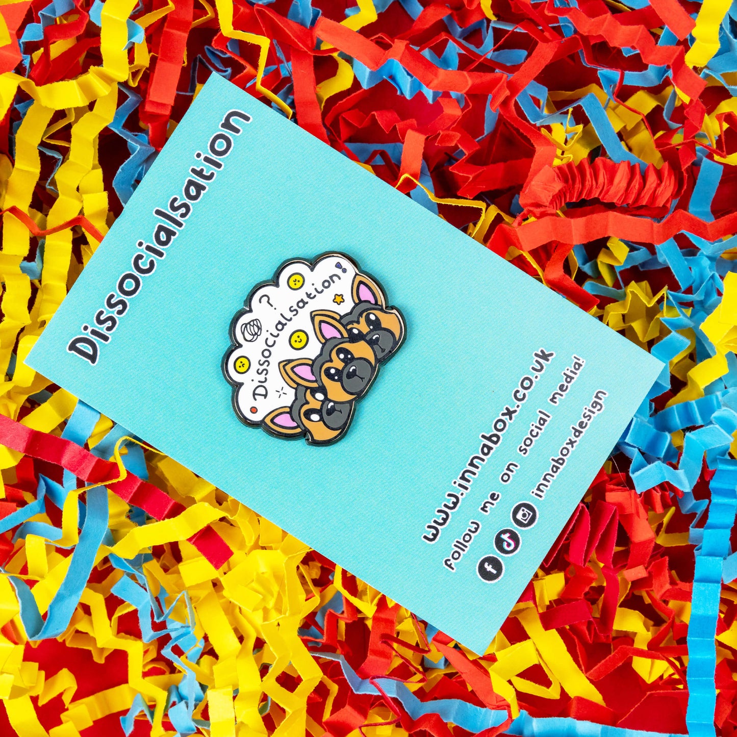 The Dissocialsation Enamel Pin - Dissociation on blue backing card on a red, blue and yellow card confetti background. Three confused brown and black Alsatian dog heads with their ears perked up, above them is a white cloud with sad and happy yellow faces, sparkles, question marks and black text reading 'dissocialsation!'. The design is raising awareness for dissociation.