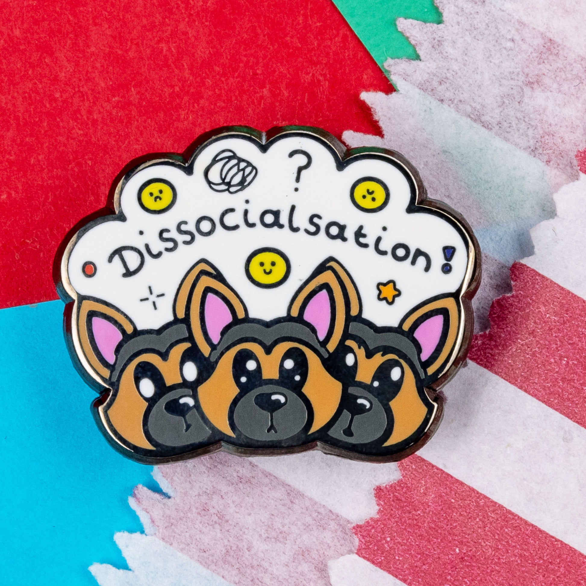 The Dissocialsation Enamel Pin - Dissociation on a red, teal and blue background next to a red and white striped paper bag. Three confused brown and black Alsatian dog heads with their ears perked up, above them is a white cloud with sad and happy yellow faces, sparkles, question marks and black text reading 'dissocialsation!'. The design is raising awareness for dissociation.