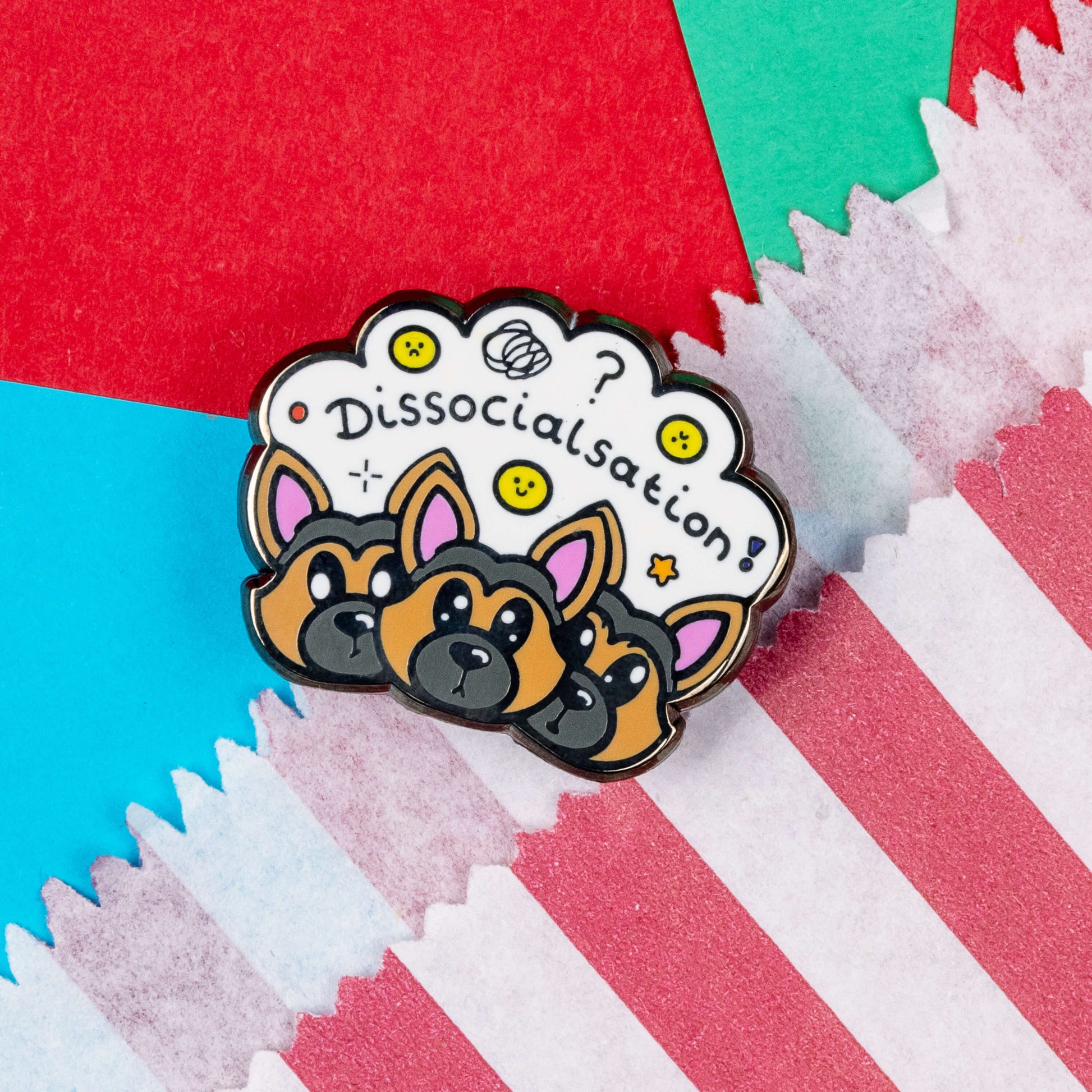 The Dissocialsation Enamel Pin - Dissociation on a red, teal and blue background next to a red and white striped paper bag. Three confused brown and black Alsatian dog heads with their ears perked up, above them is a white cloud with sad and happy yellow faces, sparkles, question marks and black text reading 'dissocialsation!'. The design is raising awareness for dissociation.