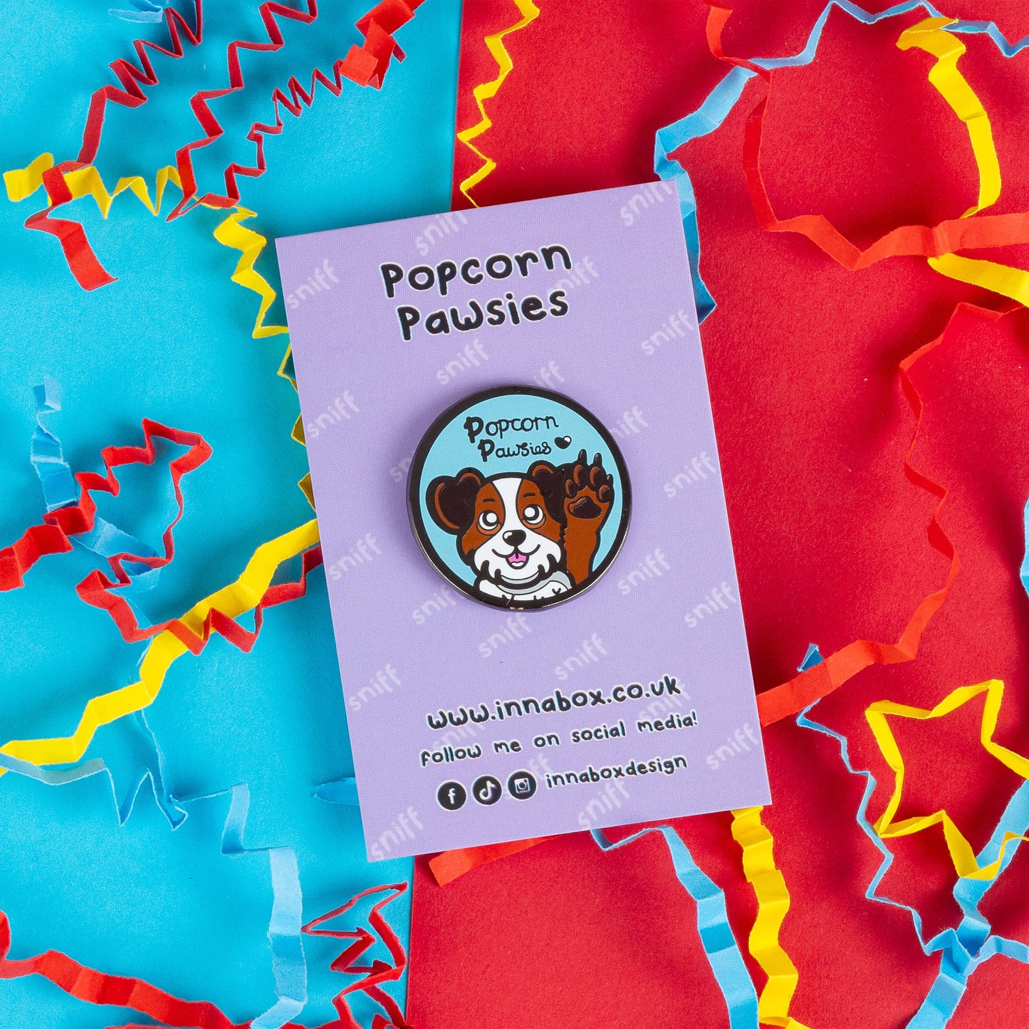 Innabox enamel pin featuring a cute dog with one paw raised, as if waving, on a lavender backing card that reads "Popcorn Pawsies." The dog's expression is cheerful, with brown and white fur, against a light blue background. Surrounded by vibrant red, blue, and yellow paper confetti, this playful pin celebrates the adorable smell of dog paws. 