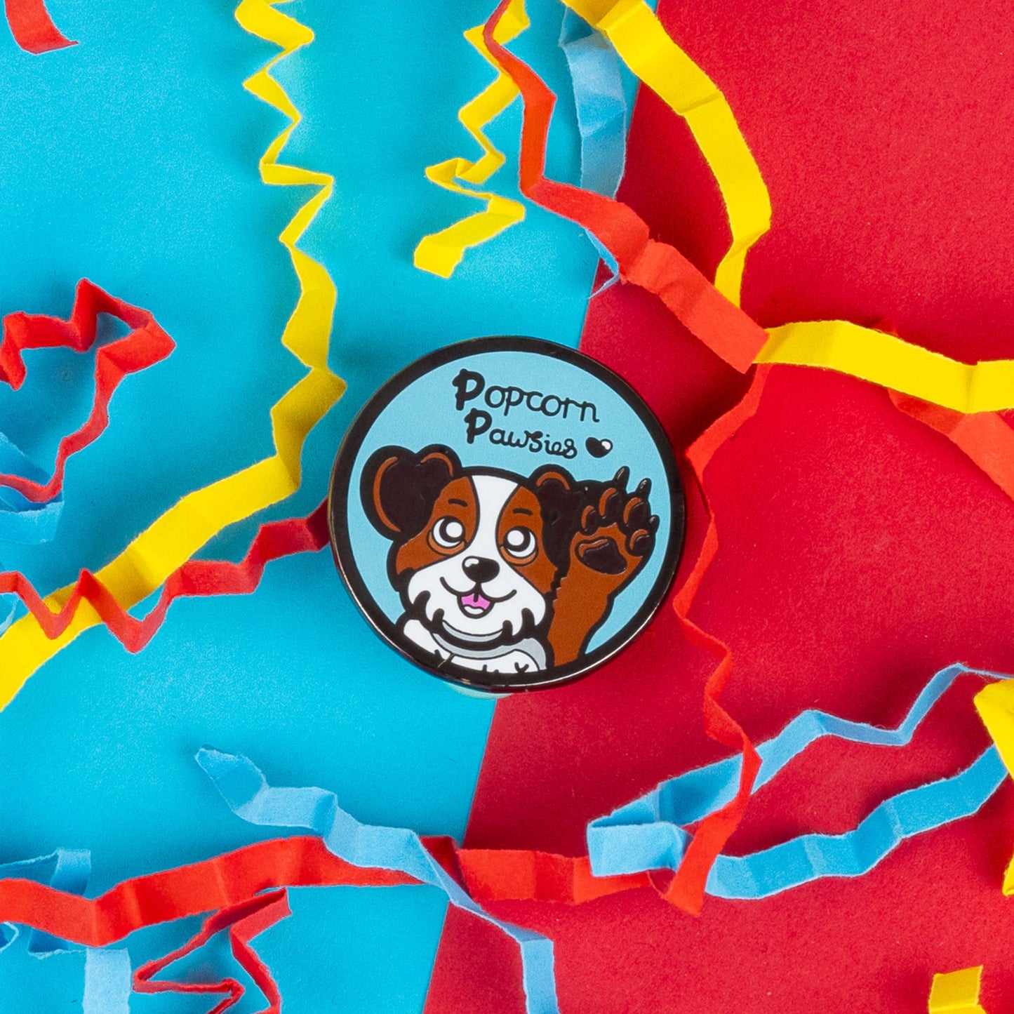 Close-up of Innabox's "Popcorn Pawsies" enamel pin, featuring a cartoon dog waving, with a happy expression on its face. The pin shows the dog in brown and white colours with the text "Popcorn Pawsies" above it. Surrounded by colourful confetti, this pin is an ideal accessory for dog lovers who appreciate fun and unique designs celebrating the love of dogs.