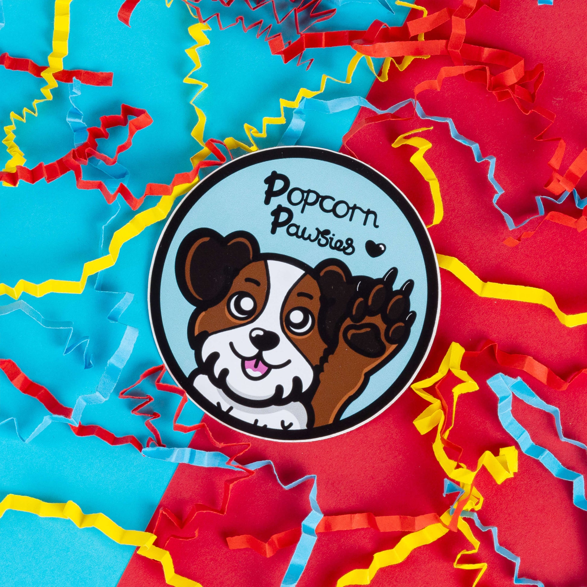 Innabox sticker featuring a cheerful cartoon dog with one paw raised, as if waving. The design includes the text 'Popcorn Pawsies' with a small heart, referencing the cute smell of a dog’s paws. The dog has a happy expression, with brown and white fur, surrounded by vibrant red, blue, and yellow paper confetti.