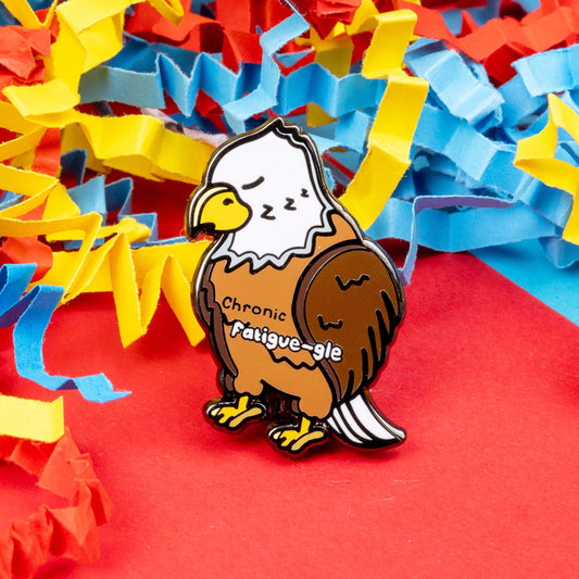 The Chronic Fatigue-le Enamel Pin - Chronic Fatigue on a red, blue and yellow card confetti background. The enamel pin is a sleeping tired brown and white eagle with a yellow beak and feet, across the middle in black text reads 'chronic' and fatigue-gle' in white text. The design was created to raise awareness for chronic fatigue / ME / CFS.