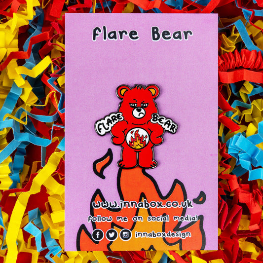 Flare Bear enamel pin on pink backing card with flames on held in front of a red, blue and yellow card confetti background. The enamel pin is of a red bear with a fed up expression and hands on its hips. There is a white circle on its belly with flames inside. Flare Bear is written on the pin. The enamel pin is designed to raise awareness for chronic illness flare ups.