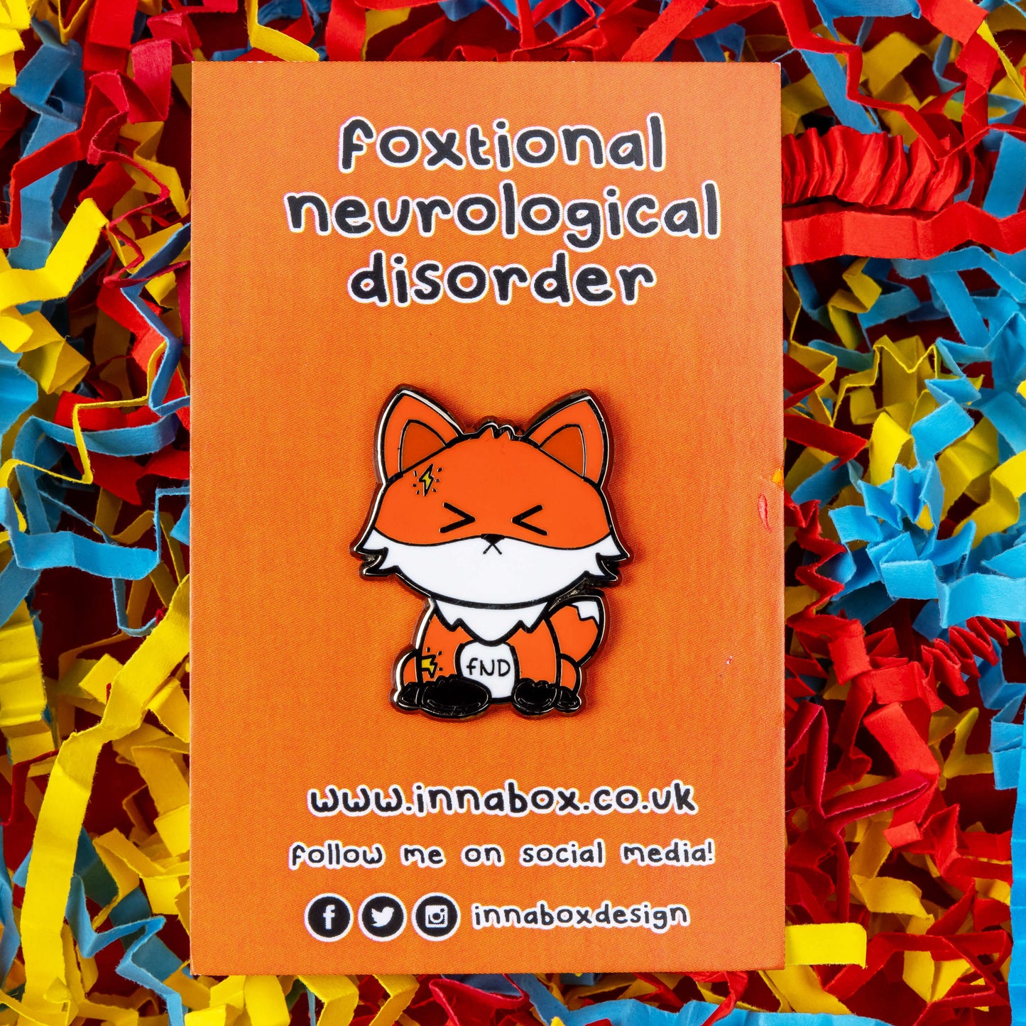 Foxtional Neurological Disorder - Functional Neurological Disorder (FND) Enamel Pin on orange backing card in front of a red, yellow and blue coloured card background. The enamel pin is a fox with a pained expression on its face and yellow lightning illustration on its head and arm. The enamel pin is designed to raise awareness for Functional Neurological Disorder (FND)