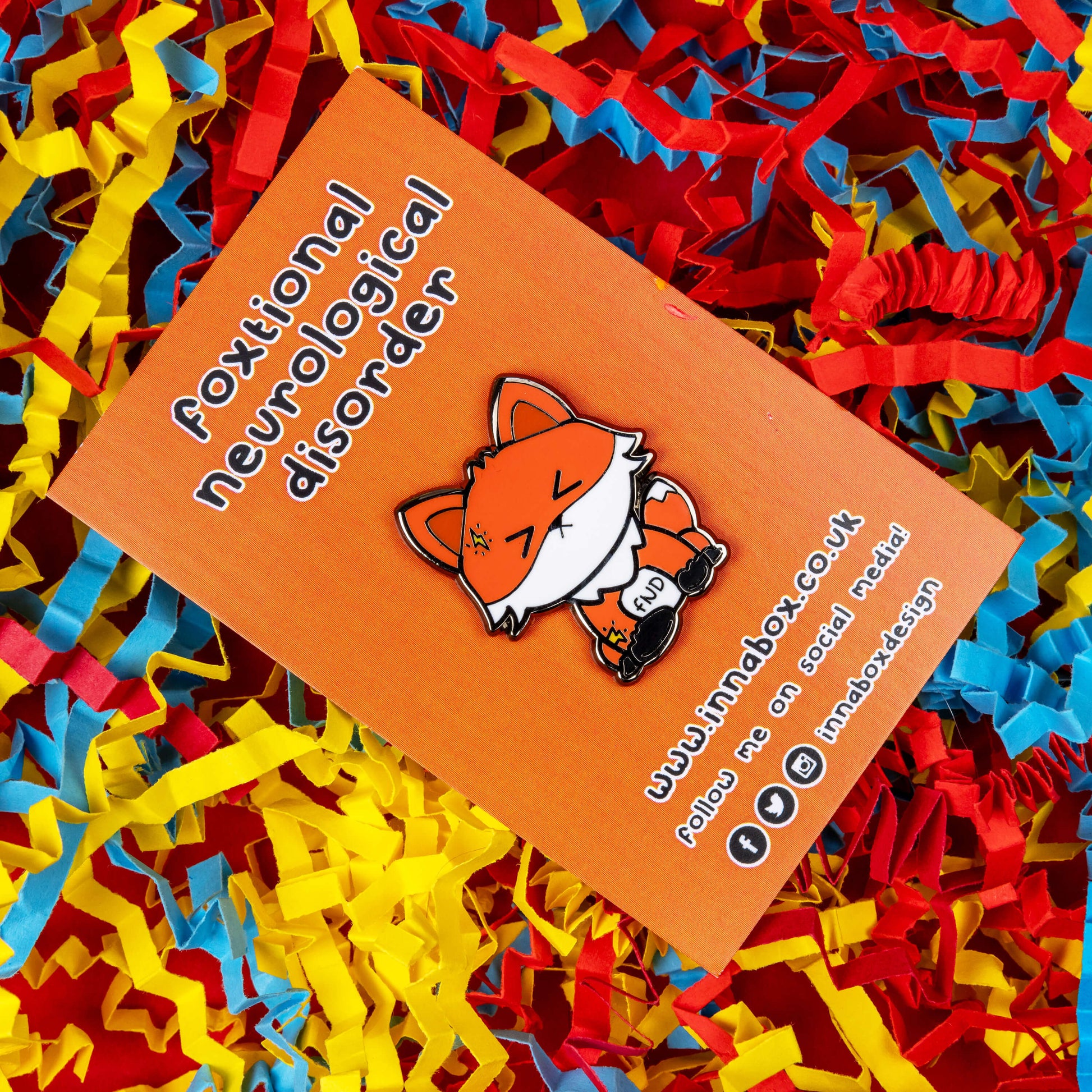 Foxtional Neurological Disorder - Functional Neurological Disorder (FND) Enamel Pin on orange backing card in front of a red, yellow and blue coloured card background. The enamel pin is a fox with a pained expression on its face and yellow lightning illustration on its head and arm. The enamel pin is designed to raise awareness for Functional Neurological Disorder (FND)