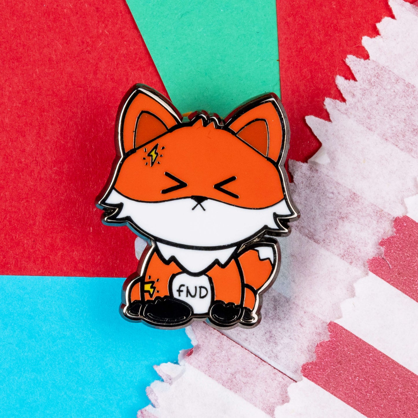 Foxtional Neurological Disorder - Functional Neurological Disorder (FND) Enamel Pin shown on a blue, red and teal background next to a red and white striped paper bag. The enamel pin is a fox with a pained expression on its face and yellow lightning illustration on its head and arm. The enamel pin is designed to raise awareness for Functional Neurological Disorder (FND)