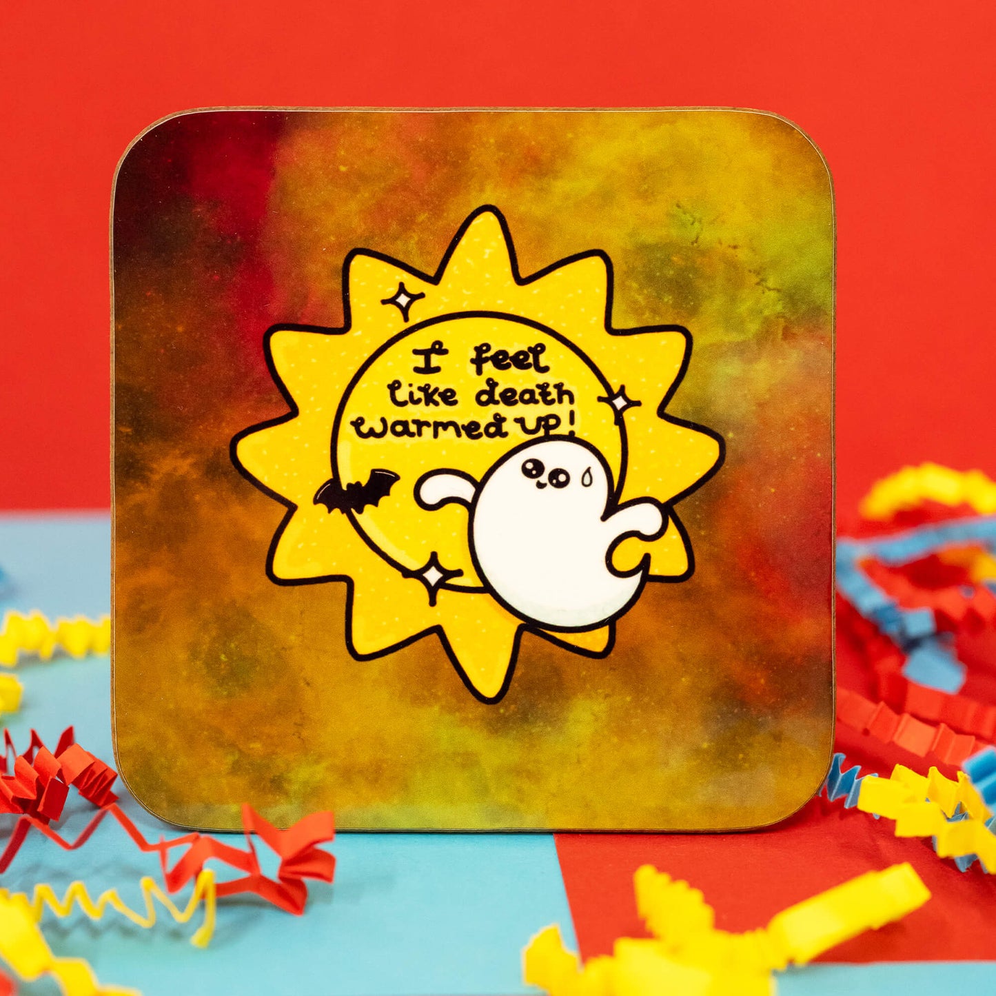 Close-up of the Death Warmed Up Coaster by Innabox. The square coaster has rounded corners and a mottled background in shades of yellow, orange, and green. The design features a worried-looking white ghost with a sweat drop, standing in front of a bright yellow sunburst with the phrase "I feel like death warmed up!" in playful black text. A tiny bat and star details surround the design. Colourful crinkle paper surrounds the coaster.