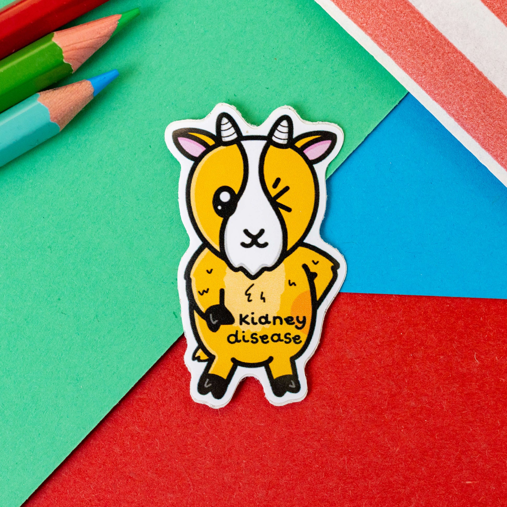 The Kidney Disease Goat Sticker on a red, blue and green background with colouring pencils and red stripe candy bag. The goat shape sticker is orange with little horns winking with a red right hand side, across its middle is black text that reads 'Kidney disease'. The hand drawn design is raising awareness for kidney diseases.