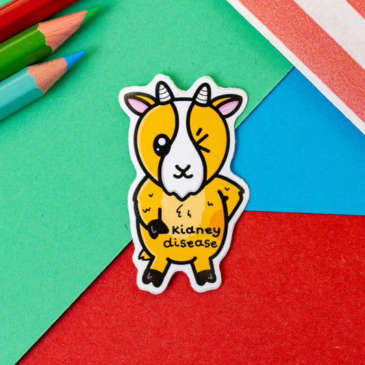 The Kidney Disease Goat Sticker on a red, blue and green background with colouring pencils and red stripe candy bag. The goat shape sticker is orange with little horns winking with a red right hand side, across its middle is black text that reads 'Kidney disease'. The hand drawn design is raising awareness for kidney diseases.