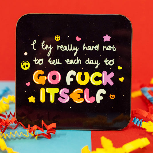 Close-up of the I Try Really Hard Coaster by Innabox. The square coaster has rounded corners and a glossy black background. The design features the phrase "I try really hard not to tell each day to GO F*** ITSELF" in playful, multicoloured bubble letters, with pink hearts, yellow smileys, stars, and dots scattered around. Colourful red, blue, and yellow crepe paper surrounds the coaster.