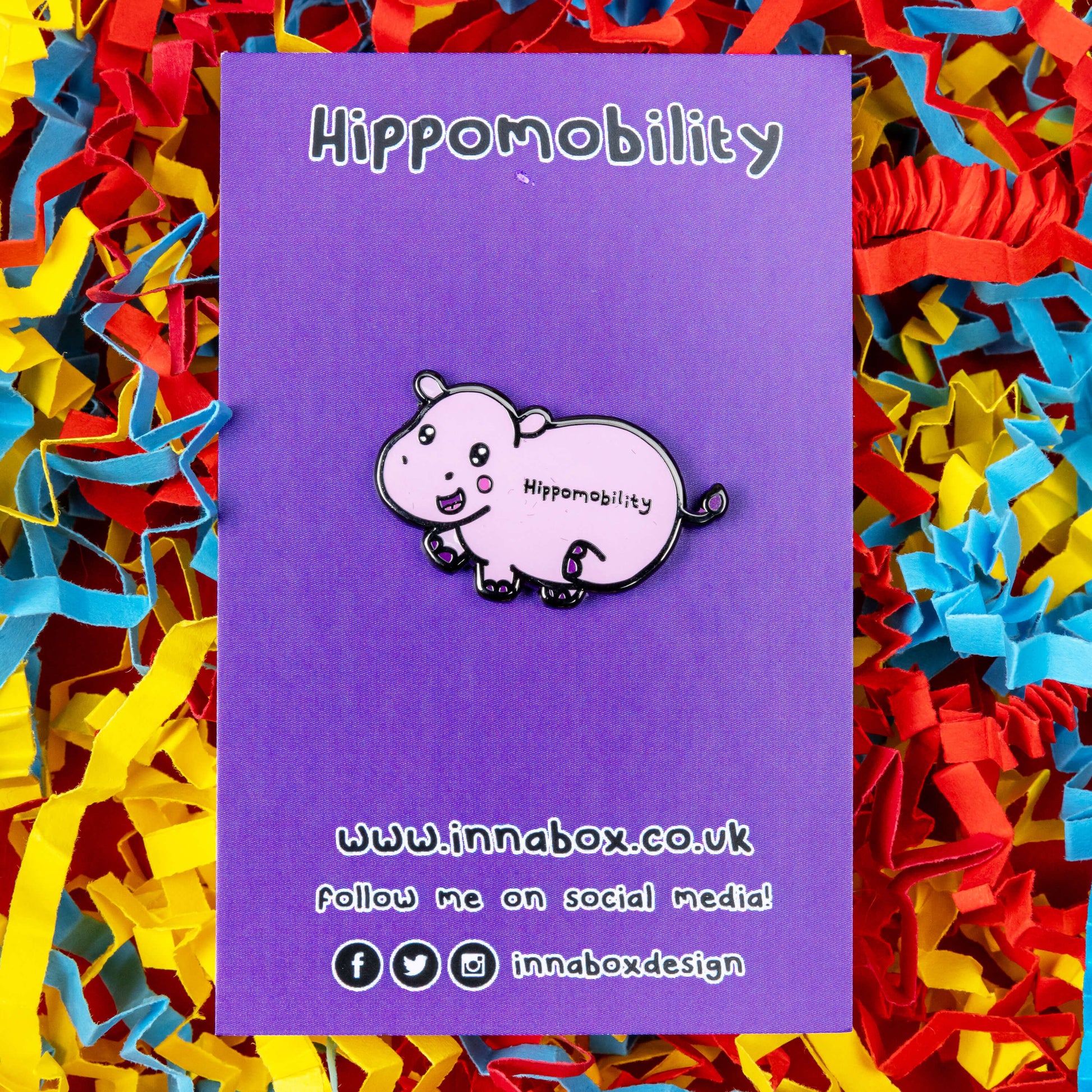 Hippmobility Enamel Pin - Hyper Mobility shown on purple backing card on a red, blue and yellow coloured card confetti background. The enamel pin is of a pink happy hippo with the text hippomobility written on its stomach. The enamel pin is designed to raise awareness for hyper mobility