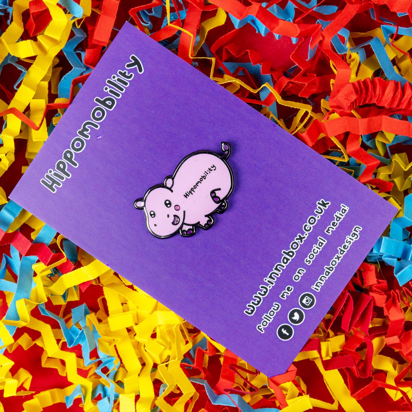 Hippmobility Enamel Pin - Hyper Mobility shown on purple backing card on a red, blue and yellow coloured card confetti background. The enamel pin is of a pink happy hippo with the text hippomobility written on its stomach. The enamel pin is designed to raise awareness for hyper mobility
