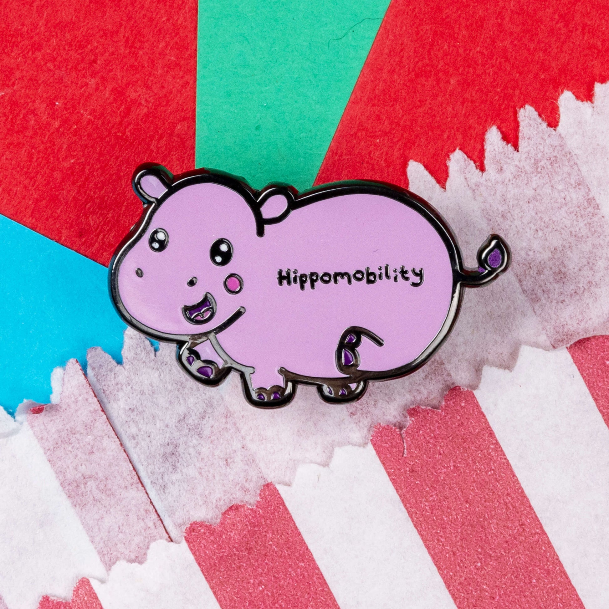 Hippmobility Enamel Pin - Hyper Mobility shown on a red and white striped paper bag on a red, teal and blue background. The enamel pin is of a pink happy hippo with the text hippomobility written on its stomach. The enamel pin is designed to raise awareness for hyper mobility
