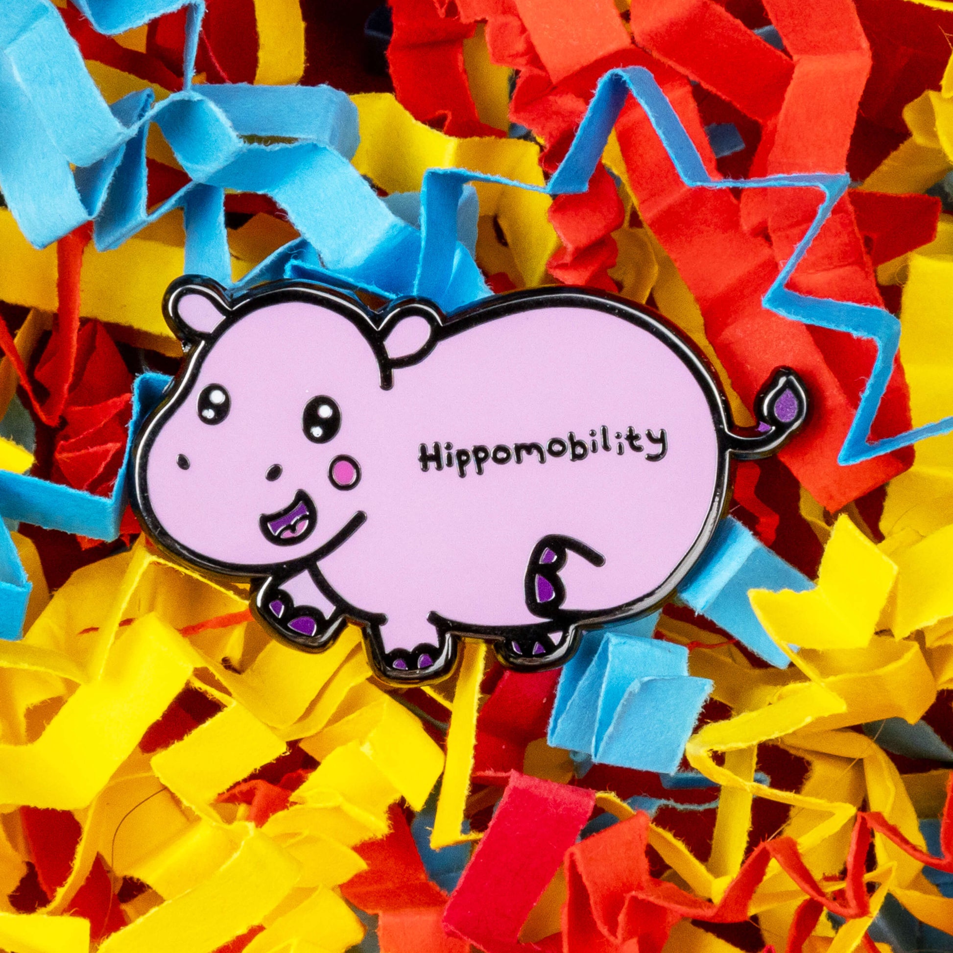 Hippmobility Enamel Pin - Hyper Mobility shown on a red, blue and yellow card confetti background. The enamel pin is of a pink happy hippo with the text hippomobility written on its stomach. The enamel pin is designed to raise awareness for hyper mobility