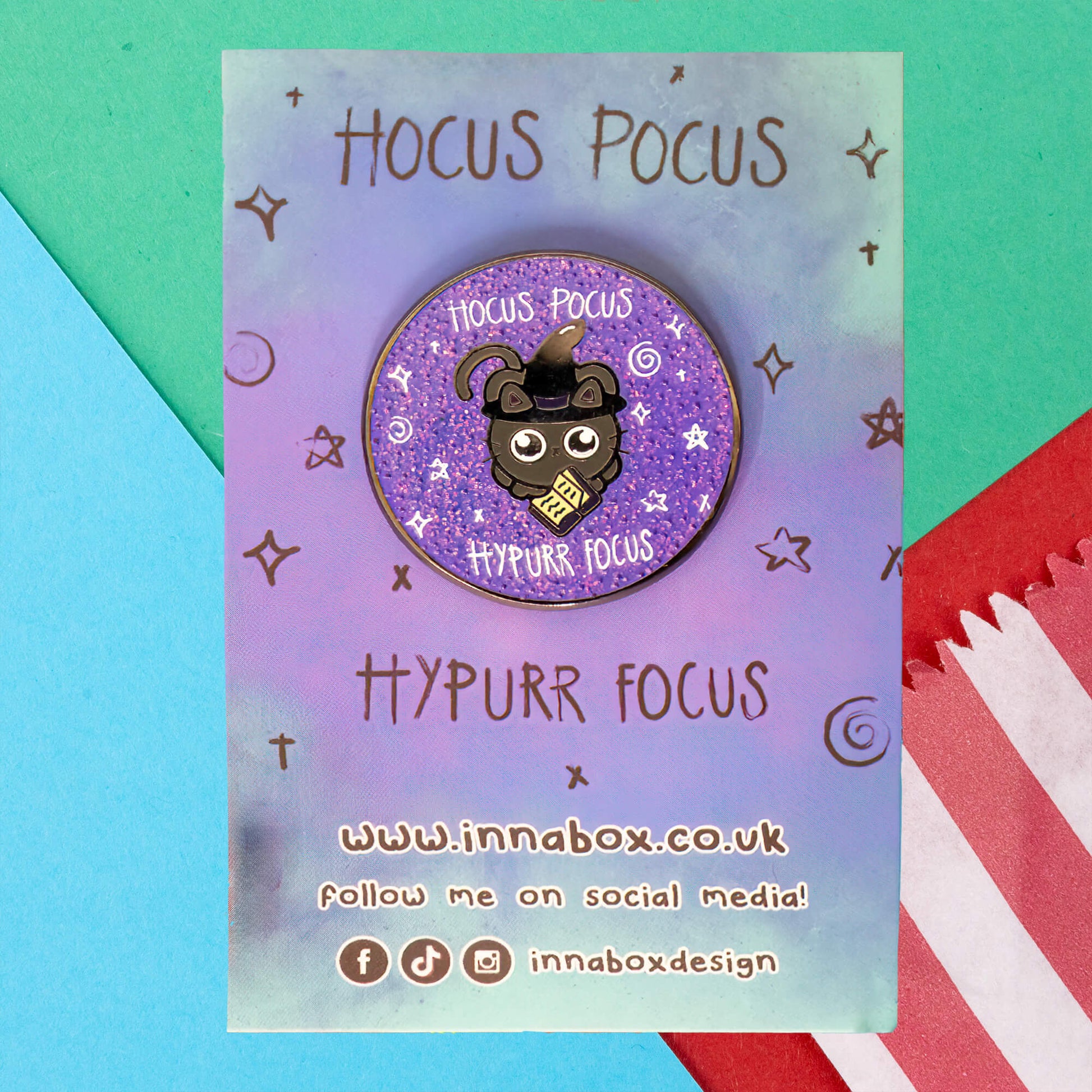 Innabox's Hocus Pocus Hypurr Focus Glittery Cat Enamel Pin on a pastel gradient backing card with star doodles. The pin shows a black cat in a witch hat holding a scroll, with the phrases "Hocus Pocus" and "Hypurr Focus" on a glittery purple background. Innabox branding and social media icons are at the bottom.