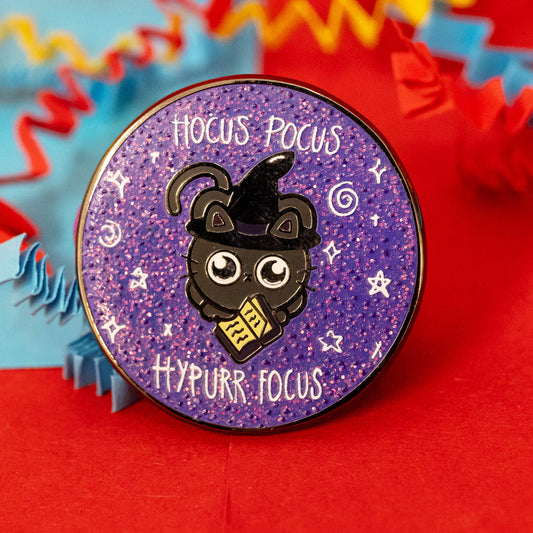 Close-up of Innabox's Hocus Pocus Hypurr Focus Glittery Cat Enamel Pin. The design features a black cat wearing a witch hat, holding a scroll, against a sparkly purple background with white stars and the phrases "Hocus Pocus" and "Hypurr Focus." The pin rests on colourful red, blue, and yellow crinkle paper.