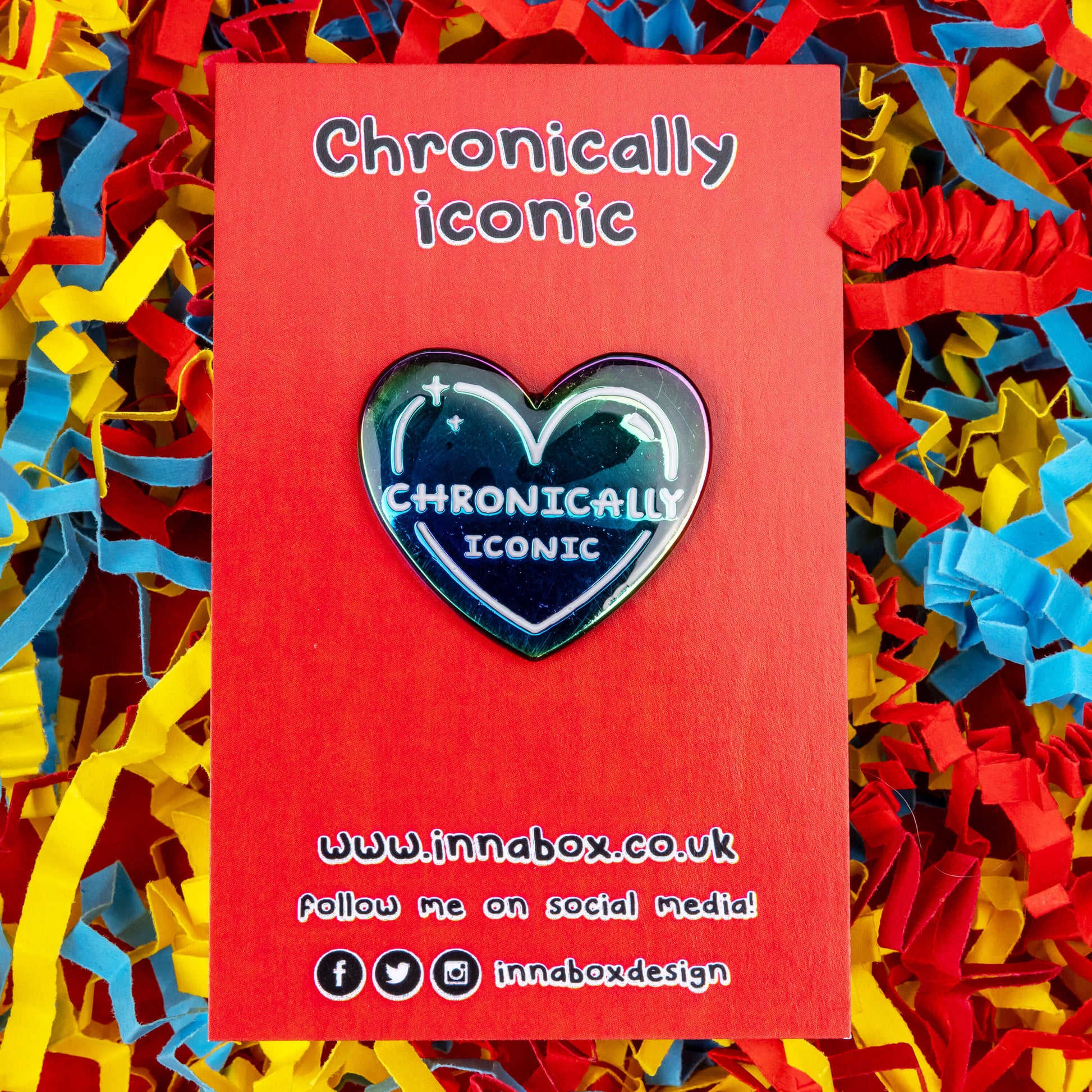 The Chronically Iconic Enamel Pin on red backing card on a red, yellow and blue card confetti background. The anodised rainbow heart shaped pin has a white outline with sparkles and text reading 'chronically iconic'. The design was created to raise awareness for chronic illness and invisible illness.