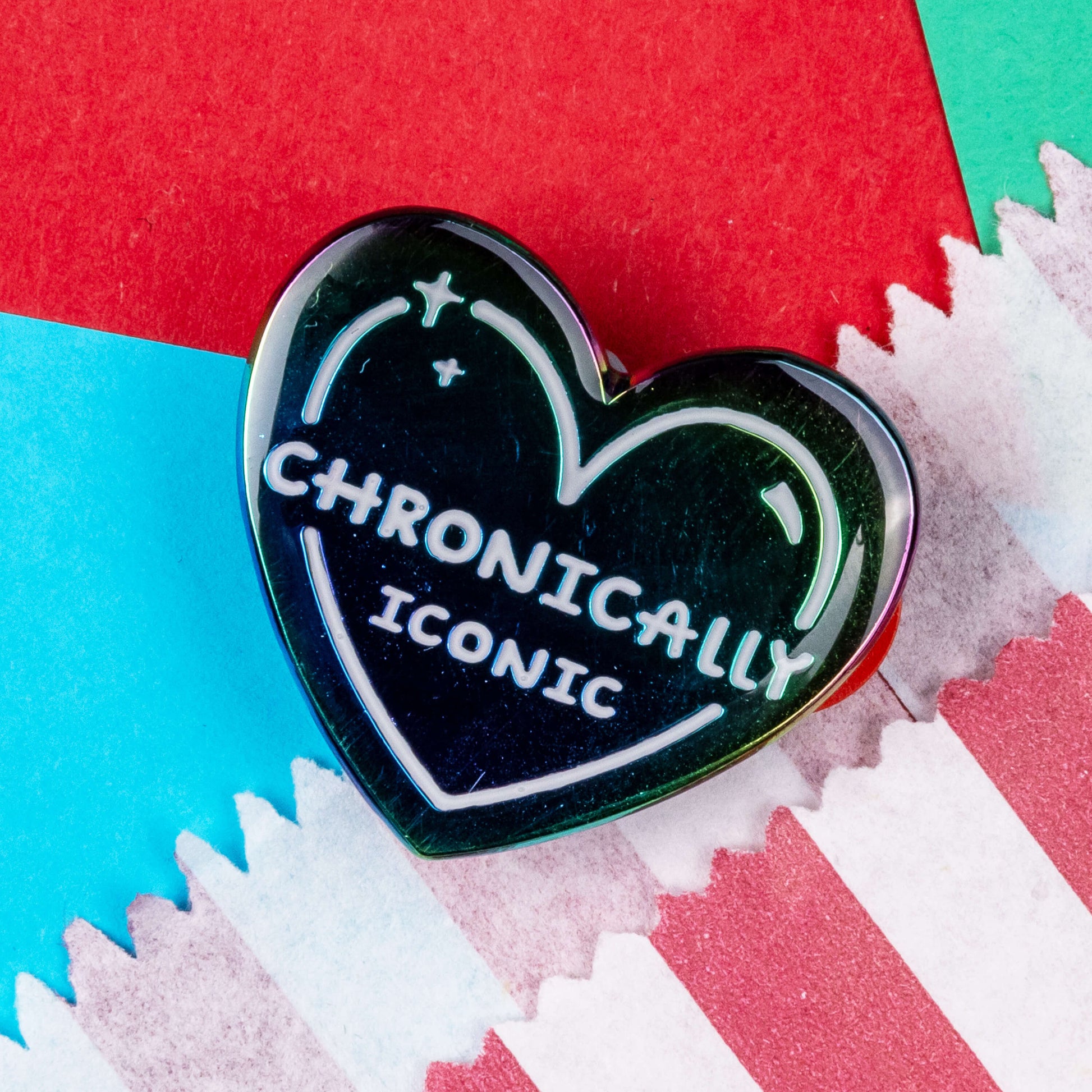The Chronically Iconic Enamel Pin on a red and blue background. The anodised rainbow heart shaped pin has a white outline with sparkles and text reading 'chronically iconic'. The design was created to raise awareness for chronic illness and invisible illness.