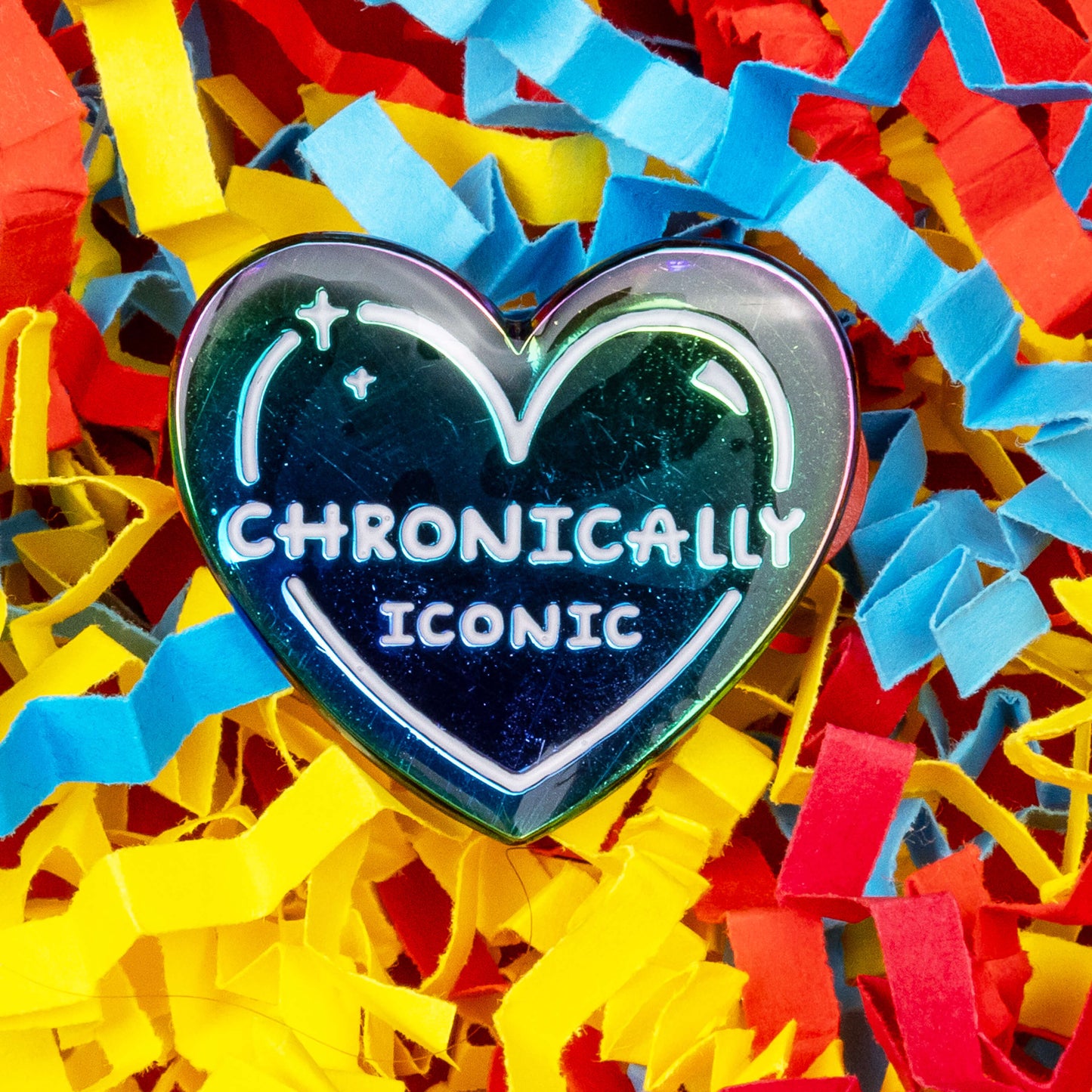 The Chronically Iconic Enamel Pin on a red, blue and yellow card confetti  background. The anodised rainbow heart shaped pin has a white outline with sparkles and text reading 'chronically iconic'. The design was created to raise awareness for chronic illness and invisible illness.