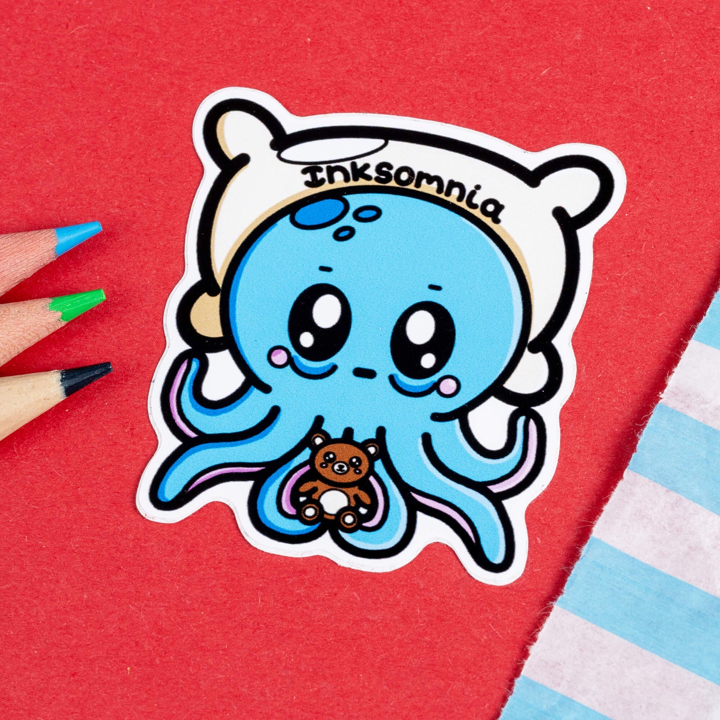 Close-up of the Inksomnia Octopus Sticker by Innabox. The sticker features a tired-looking blue octopus with big, sleepy eyes, flushed cheeks, and droopy eyelids. It’s holding a tiny brown teddy bear for comfort and wearing a white pillow-like headband with the word "Inksomnia" written across it. The background is red, with coloured pencils nearby.