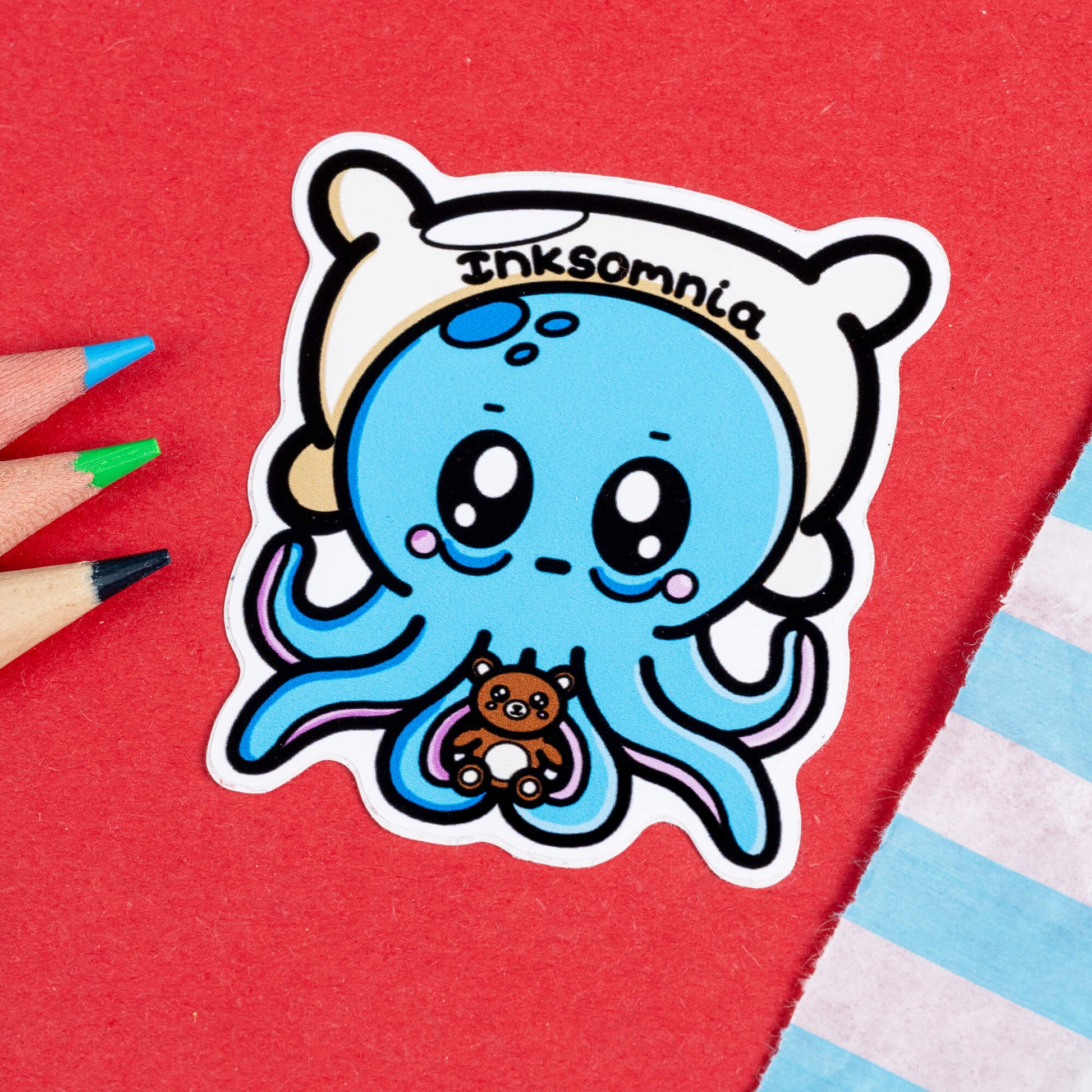 Close-up of the Inksomnia Octopus Sticker by Innabox. The sticker features a tired-looking blue octopus with big, sleepy eyes, flushed cheeks, and droopy eyelids. It’s holding a tiny brown teddy bear for comfort and wearing a white pillow-like headband with the word "Inksomnia" written across it. The background is red, with coloured pencils nearby.