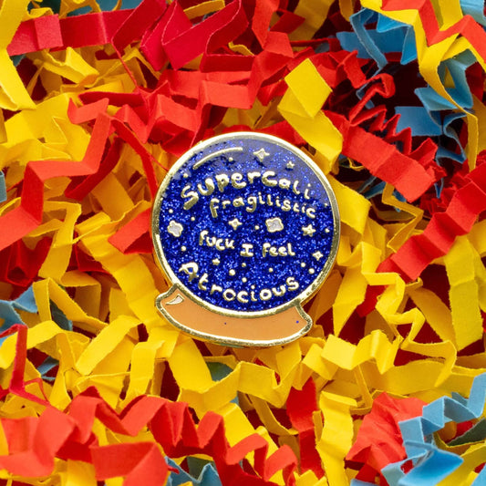 I Feel Atrocious Enamel Pin - Blue circle crystal ball shaped enamel pin with gold text reading 'supercali fragilistic fuck I feel atrocious' on. It is on a yellow, red and blue card confetti background. Enamel pin designed to raise awareness for invisible illnesses.