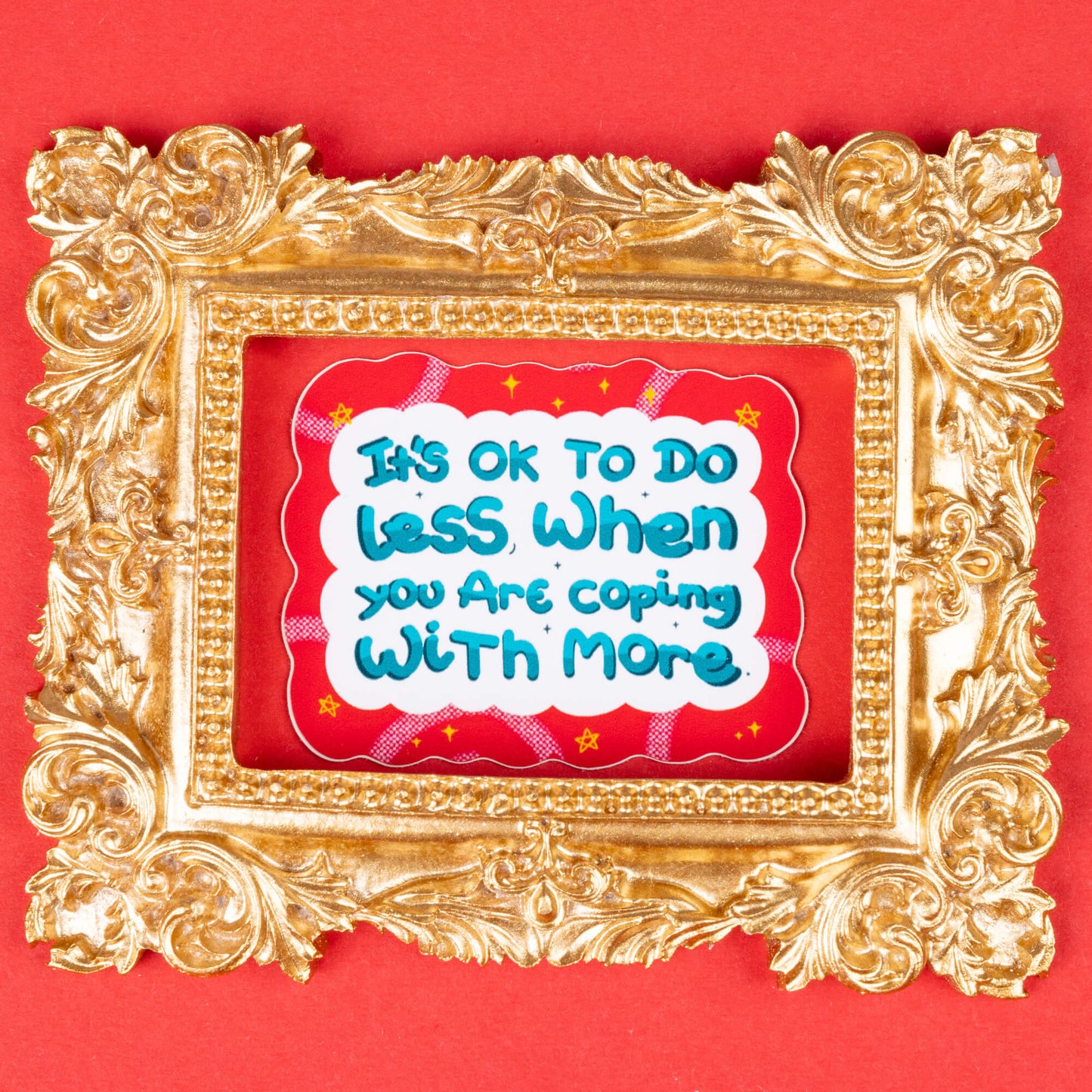 A sticker with a wavy red border and white background, displaying the text "It's OK to do less when you are coping with more" in blue and teal letters. The sticker is framed in an ornate gold picture frame and set against a red background. Hand drawn design created to raise awareness for chronic and invisible illnesses.