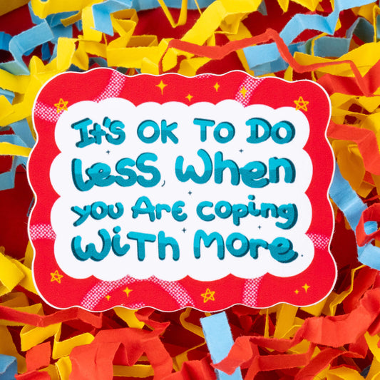 A sticker with a wavy red border and white background. The text on the sticker reads "It's OK to do less when you are coping with more" in blue and teal letters. The sticker is set against a background of colorful shredded paper in yellow, blue, and red. Hand drawn design created to raise awareness for chronic and invisible illnesses.