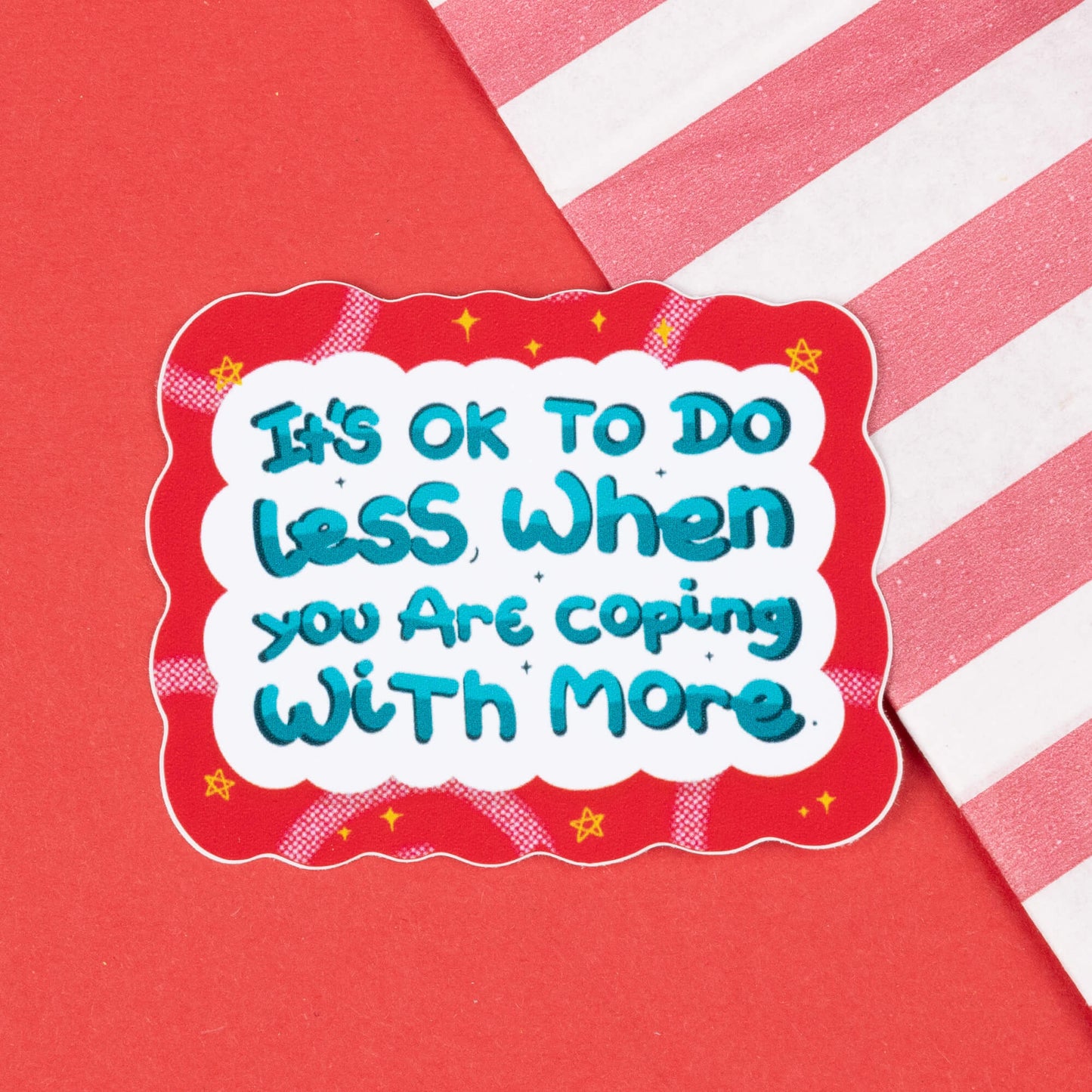 A sticker with a red wavy border and a white background featuring the text "It's OK to do less when you are coping with more" in blue and teal letters. The sticker is placed on a red background with a diagonal white and red striped pattern. Hand drawn design created to raise awareness for chronic and invisible illnesses.