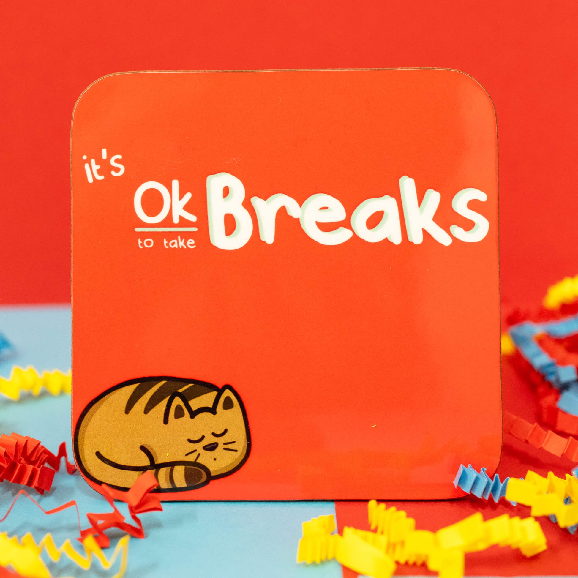 Close-up of the It's Ok To Take Breaks Cat Coaster by Innabox. The square coaster has rounded corners and a bright red background. The design features white, playful text reading "It's Ok to take Breaks," with a cute illustration of a curled-up, sleeping brown tabby cat in the bottom left corner. Colourful red, blue, and yellow crepe paper surrounds the coaster.