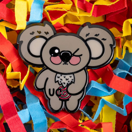 Close-up of the Ulcerative Koalitis Koala Enamel Pin by Innabox, surrounded by colourful red, yellow, and blue paper confetti. The pin features a cute koala holding a swirly object, symbolising Ulcerative Colitis.