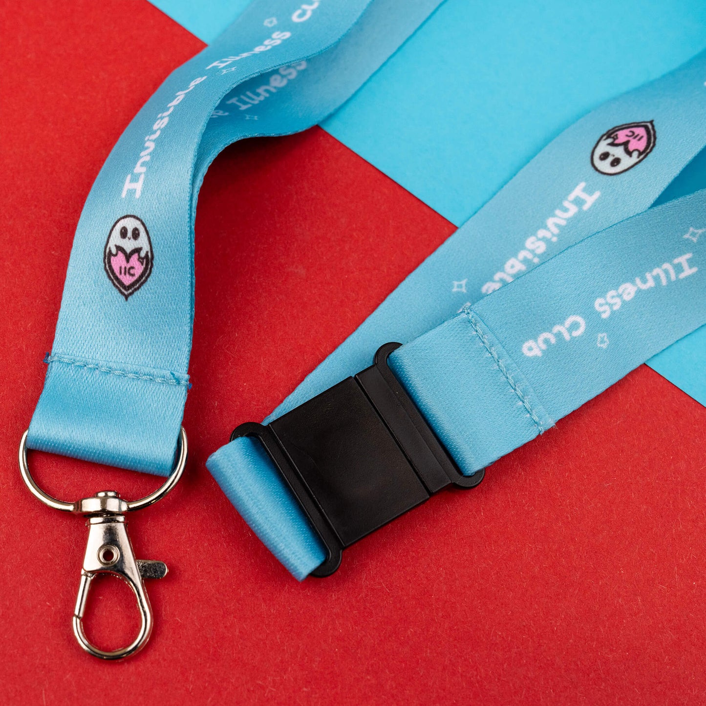 Light blue lanyard with 'Invisible Illness Club' text and a cute ghost logo, fitted with a black buckle and metal clasp. This lanyard is designed to raise awareness for invisible illnesses and is displayed against a red and blue geometric background.