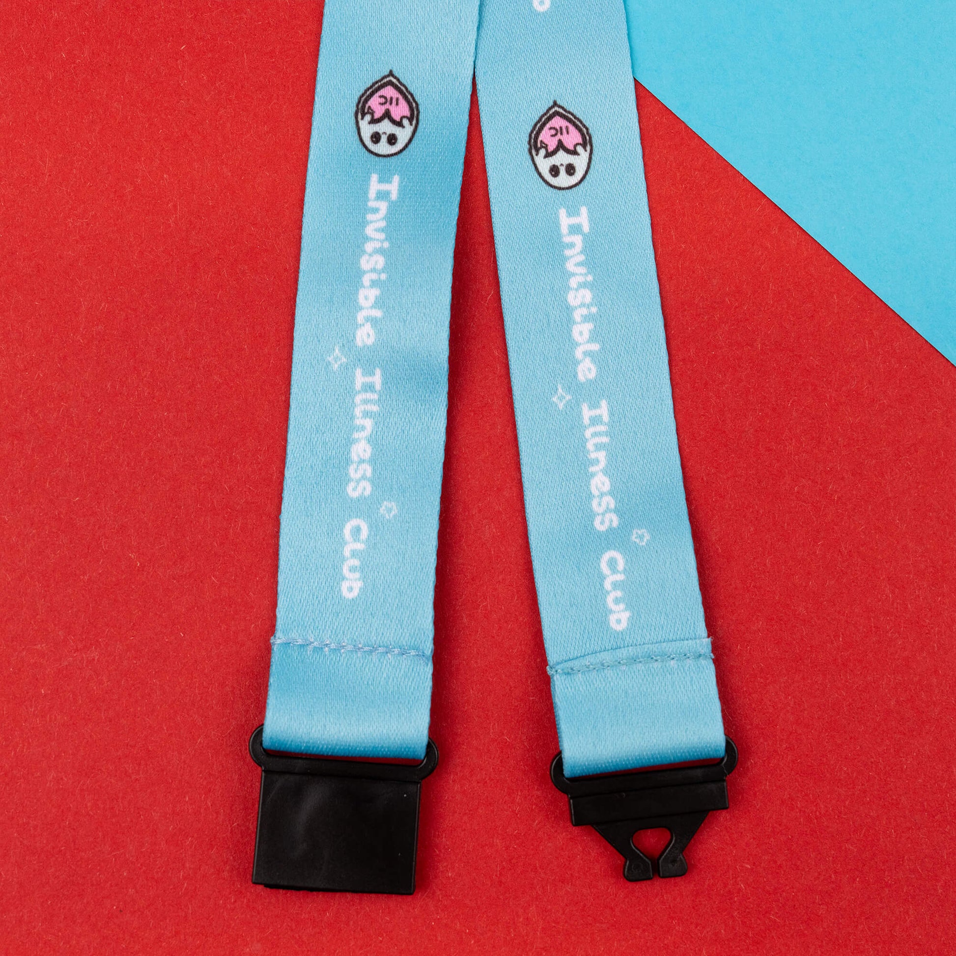 Close-up of a light blue lanyard with black safety catches, featuring the 'Invisible Illness Club' text and a whimsical ghost logo. Designed to raise awareness for invisible illnesses, it is laid out on a red and blue geometric background.