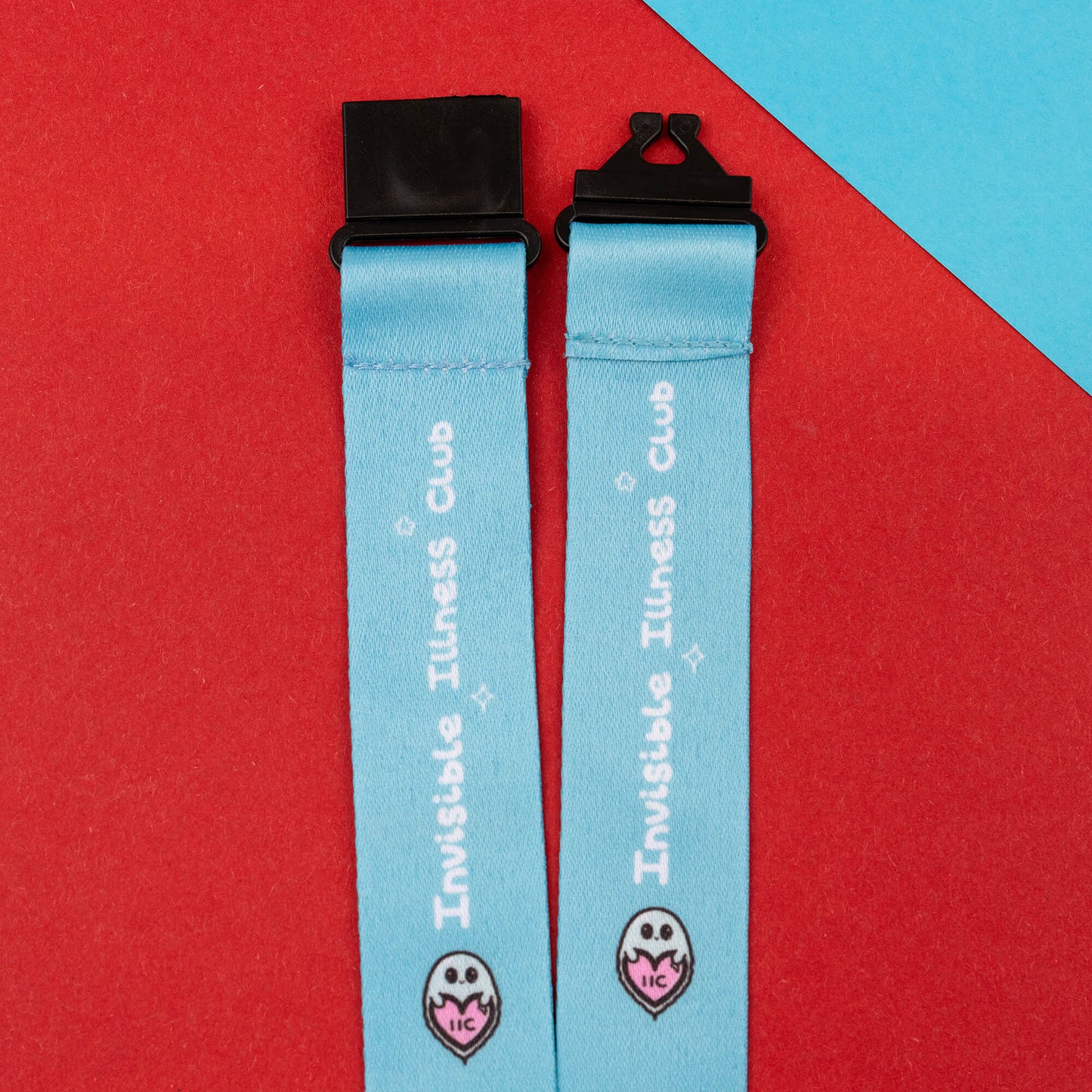 Close-up of a light blue lanyard with the 'Invisible Illness Club' text and a charming ghost logo. Equipped with black safety catches, this lanyard is designed to raise awareness for invisible illnesses and is shown on a red and blue geometric background.