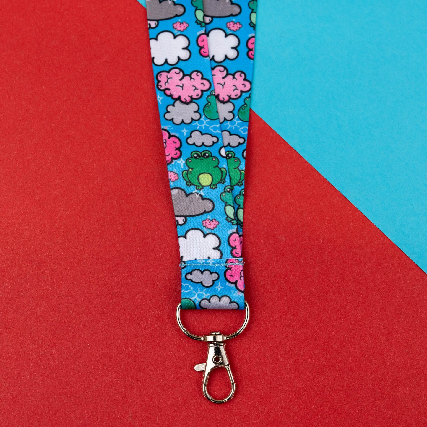 Close-up of a brain fog awareness lanyard with a metal clasp, showcasing a whimsical brain frog design with green frogs, pink brains, and white clouds on a blue background. The lanyard is equipped with black safety catches and displayed on a red and blue geometric background.