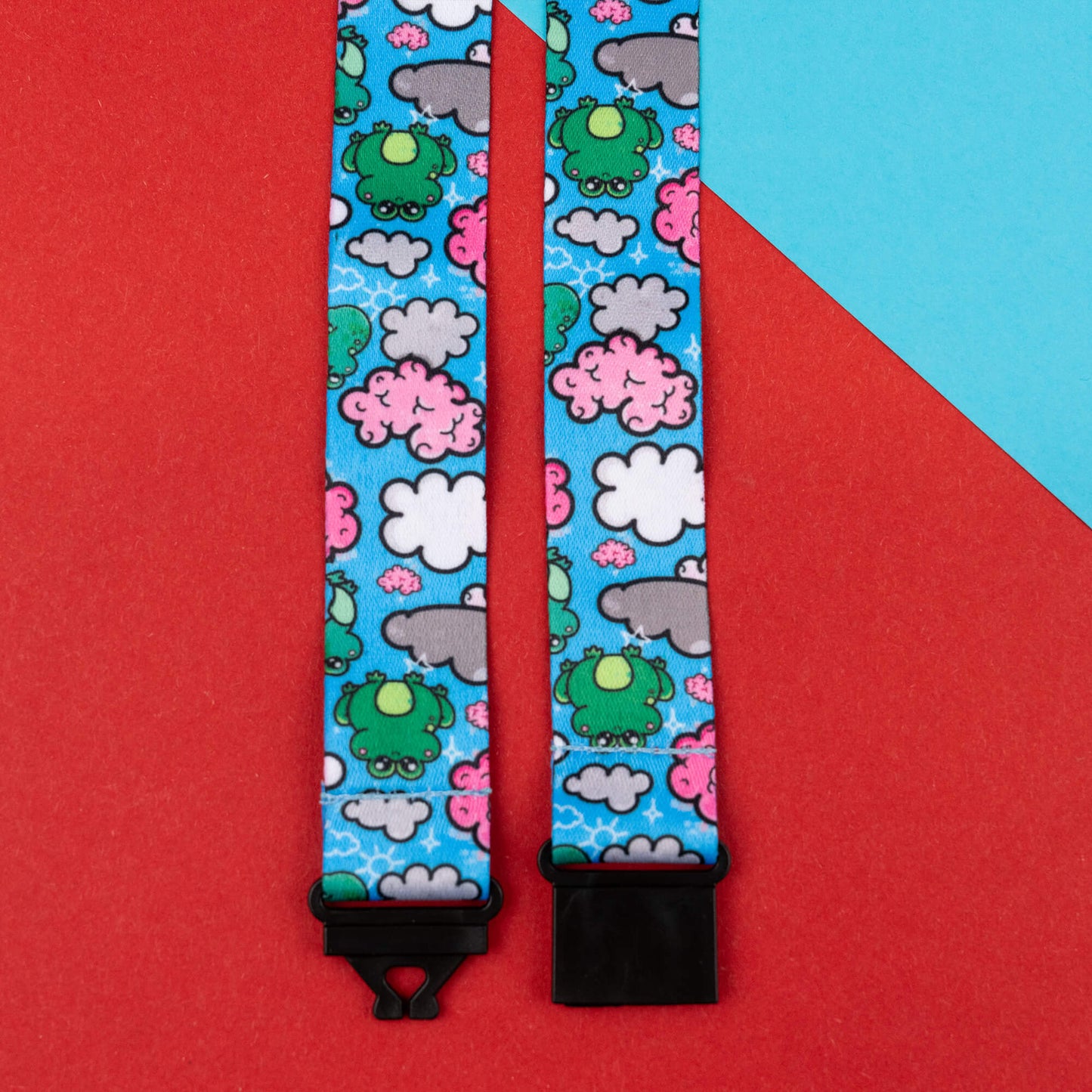 Close-up of a lanyard designed to raise awareness for brain fog, featuring a cute and playful brain frog pattern with green frogs, pink brains, and white clouds on a blue background. The lanyard is fitted with black safety catches and is laid out on a red and blue geometric background.