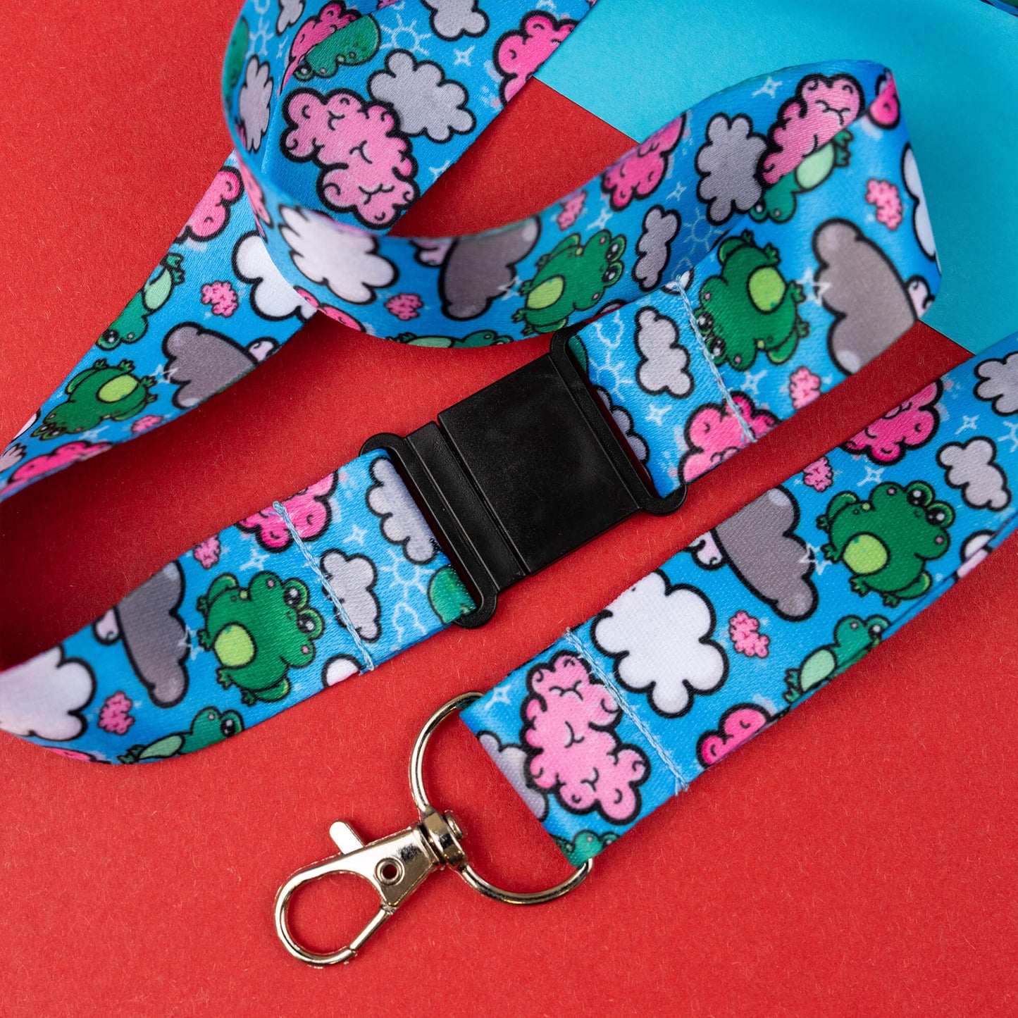 Lanyard with black buckle and metal clasp, featuring a fun and quirky brain frog pattern to raise awareness for brain fog. The design includes green frogs, pink brains, and white clouds on a blue background. The lanyard is shown against a red and blue geometric background.