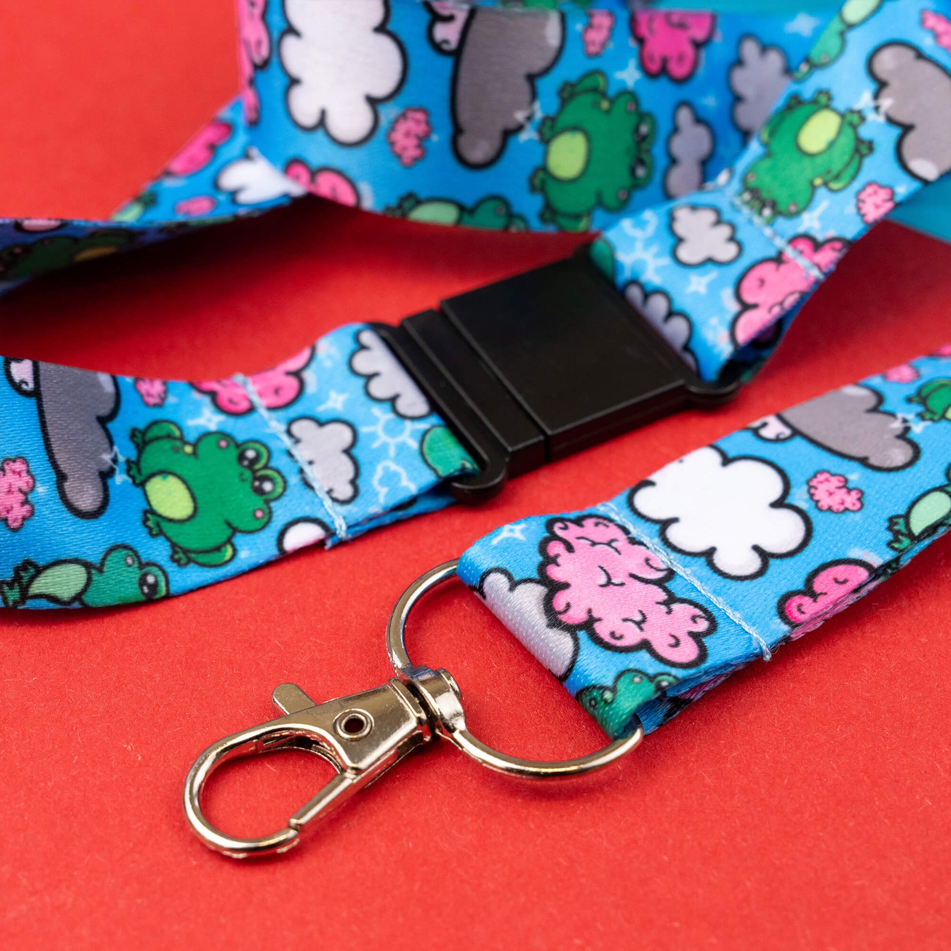 Close-up of a lanyard with a black buckle and metal clasp, decorated with a playful brain frog pattern including green frogs, pink brains, and white clouds on a blue background. Designed to raise awareness for brain fog, the lanyard is displayed on a red and blue geometric background.