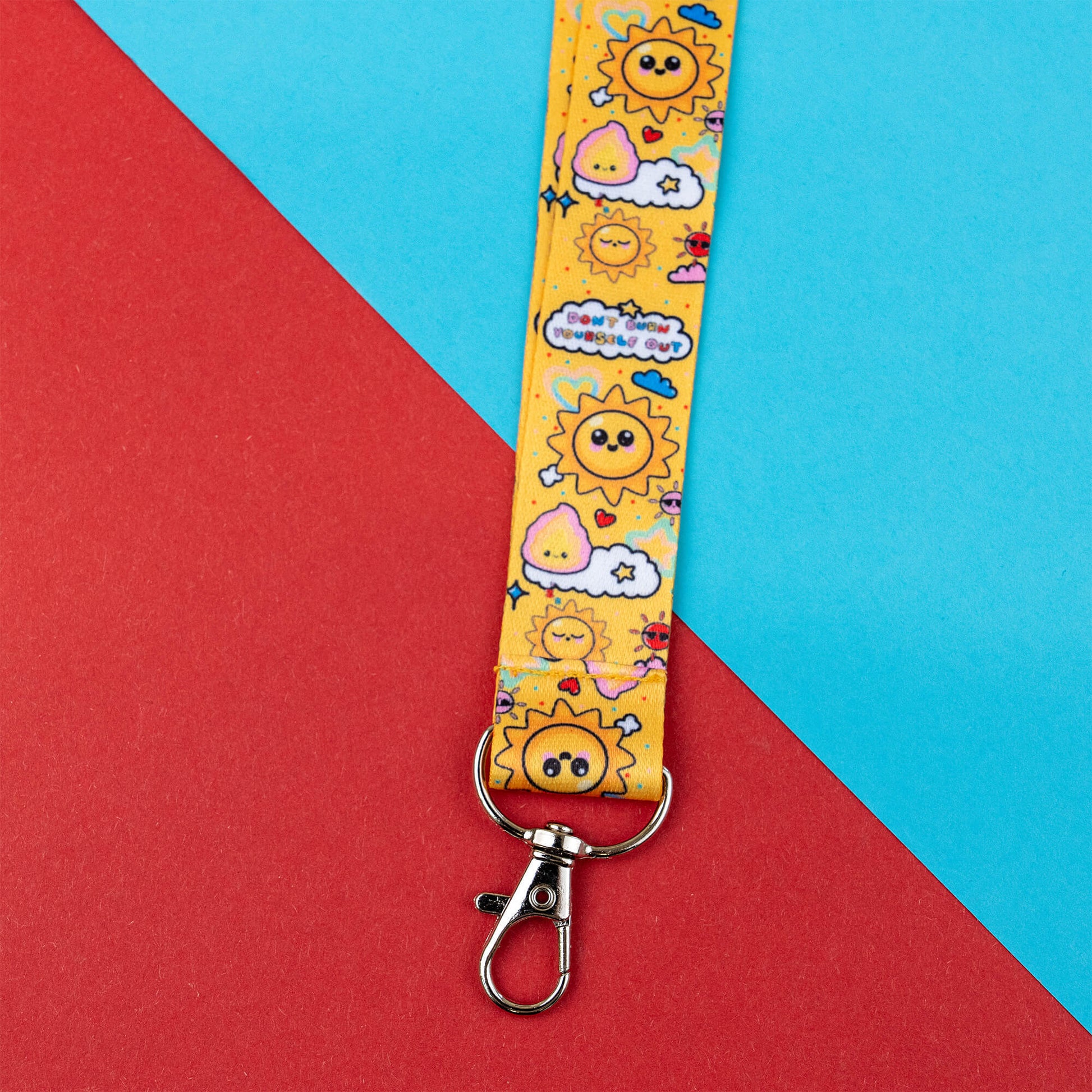 Lanyard with a cheerful sun, clouds, and motivational phrases reminding not to burn out. Yellow design promotes awareness for invisible illnesses and hidden disabilities. Features a metal clasp. Displayed on a red and blue geometric background.