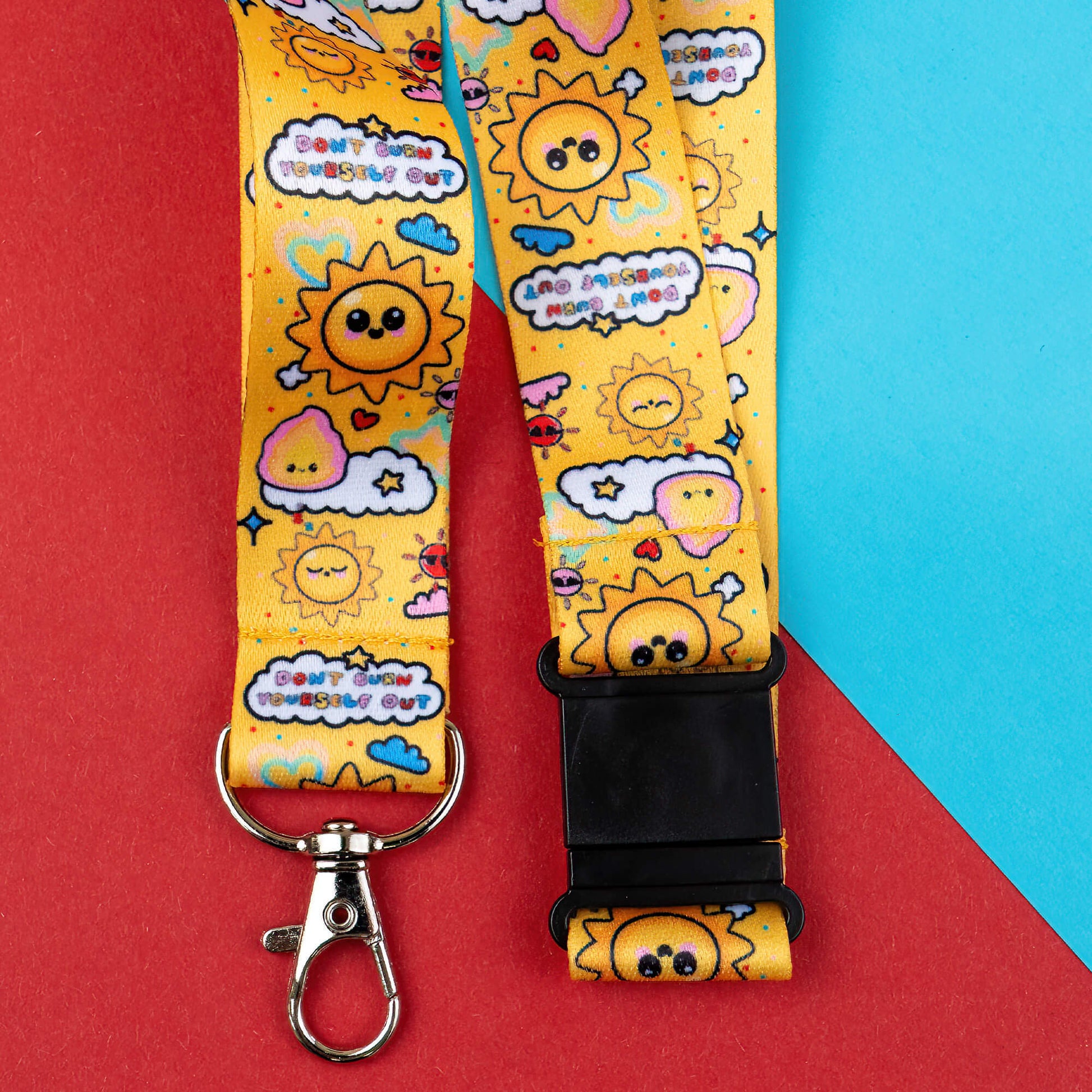 Yellow lanyard with motivational phrases and a cheerful sun design, reminding not to burn out. Raises awareness for invisible illnesses and hidden disabilities. Includes a black safety buckle and metal clasp. Shown on a red and blue geometric background.