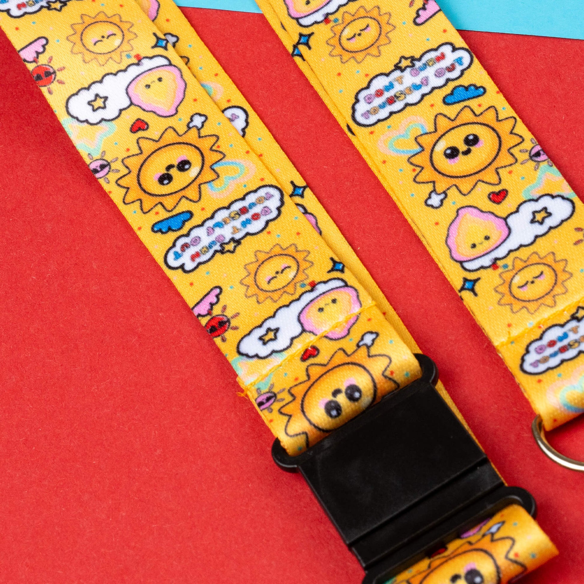 Close-up of a yellow lanyard with cheerful sun and cloud designs, including motivational phrases about not burning out. Raises awareness for invisible illnesses and hidden disabilities. Includes black safety catches. Shown on a red and blue geometric background.